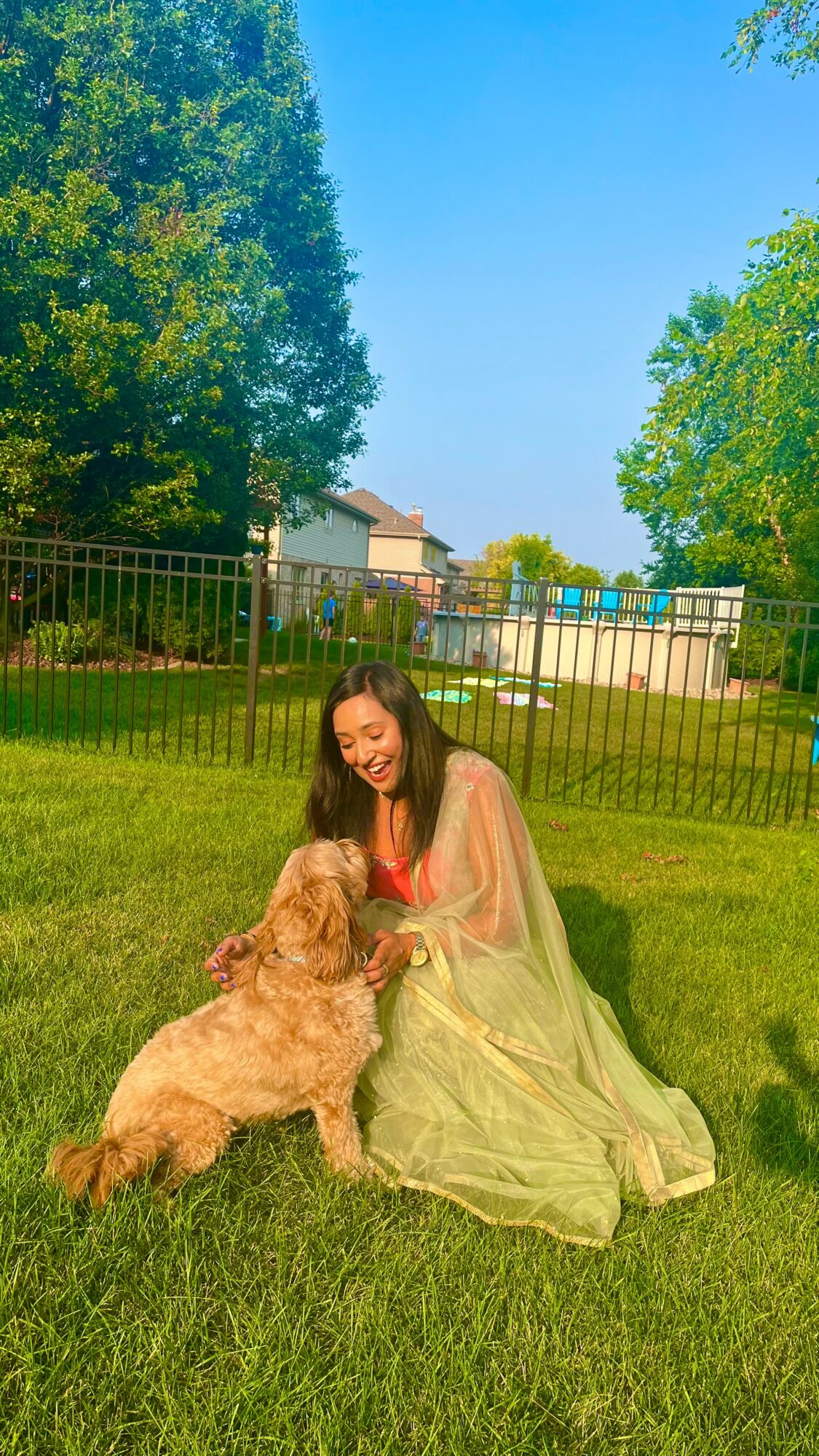 Woman in a light green dress sitting on grass with a golden retriever outdoors, trees and a fence in background.