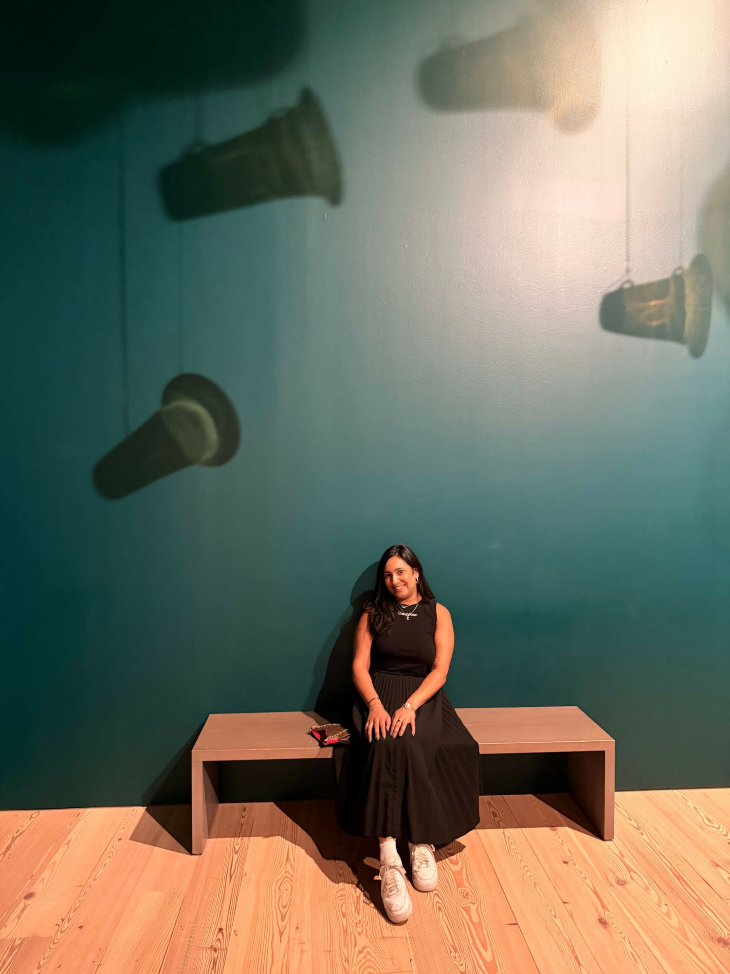 Person sitting on a bench in front of a blue wall with floating plant pots, wooden floor, and a person with long dark hair.