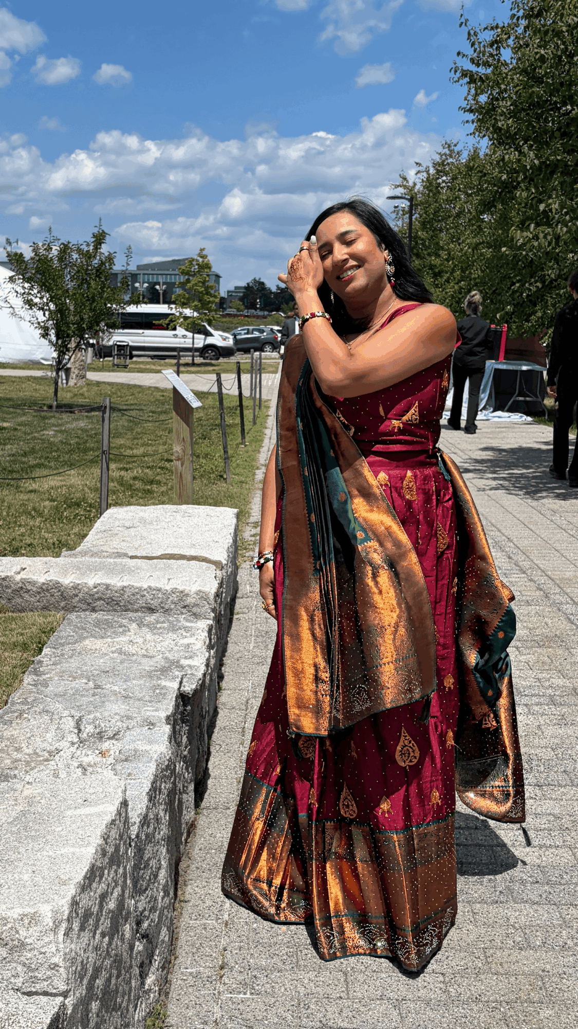 Woman in colorful traditional dress smiling outdoors, with trees and a blue sky in background.