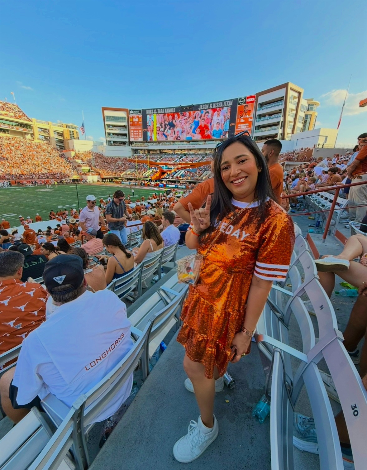 Smiling woman in orange sports jersey and white sneakers at stadium, holding a drink, with crowd and large screen in background.