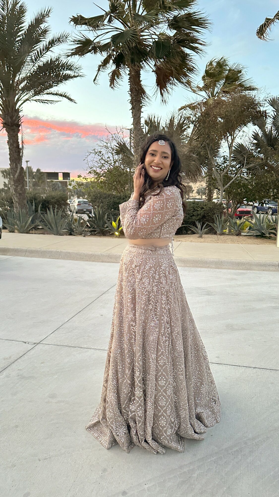Woman in a long, lace dress standing outdoors with palm trees and a sunset sky in background.