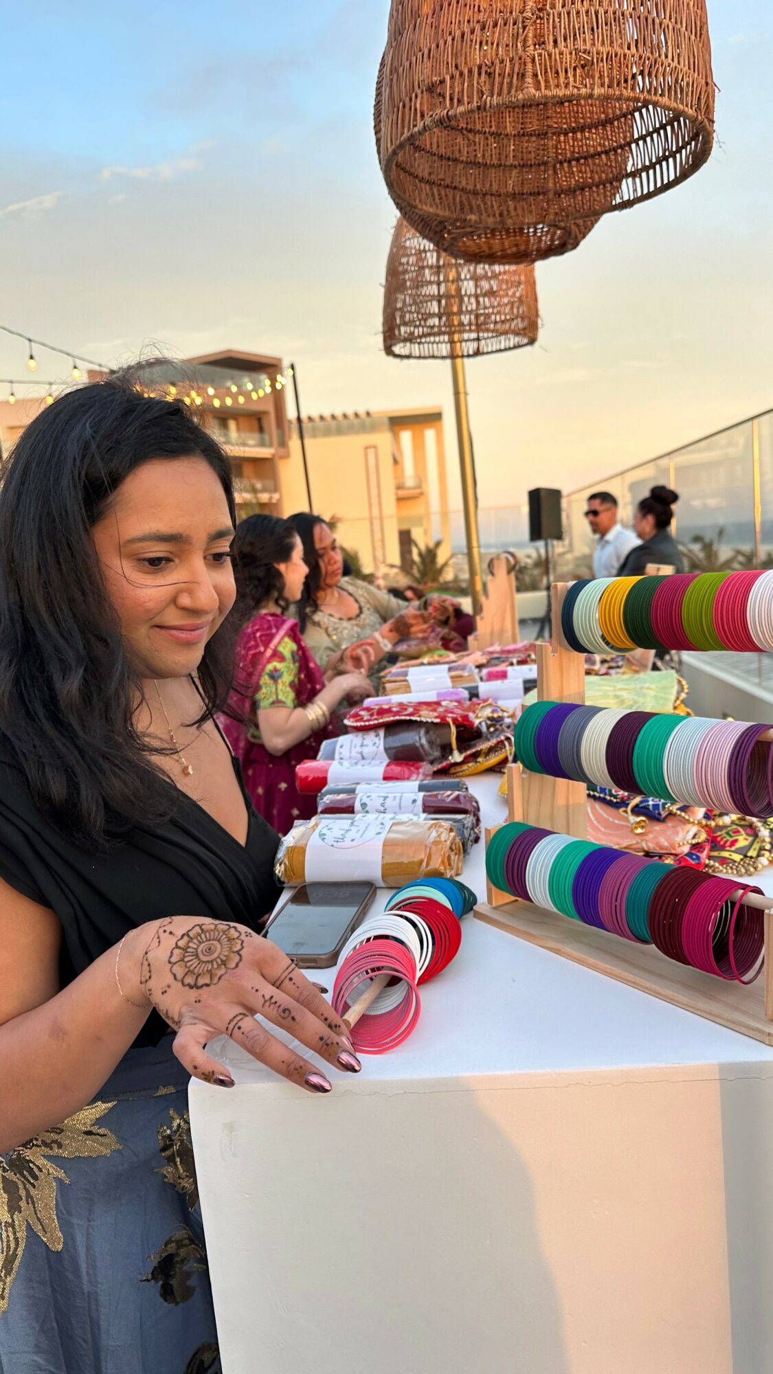 Woman with henna on her hand at an outdoor market stall with colorful rolled fabrics and woven lamps, sunset sky.