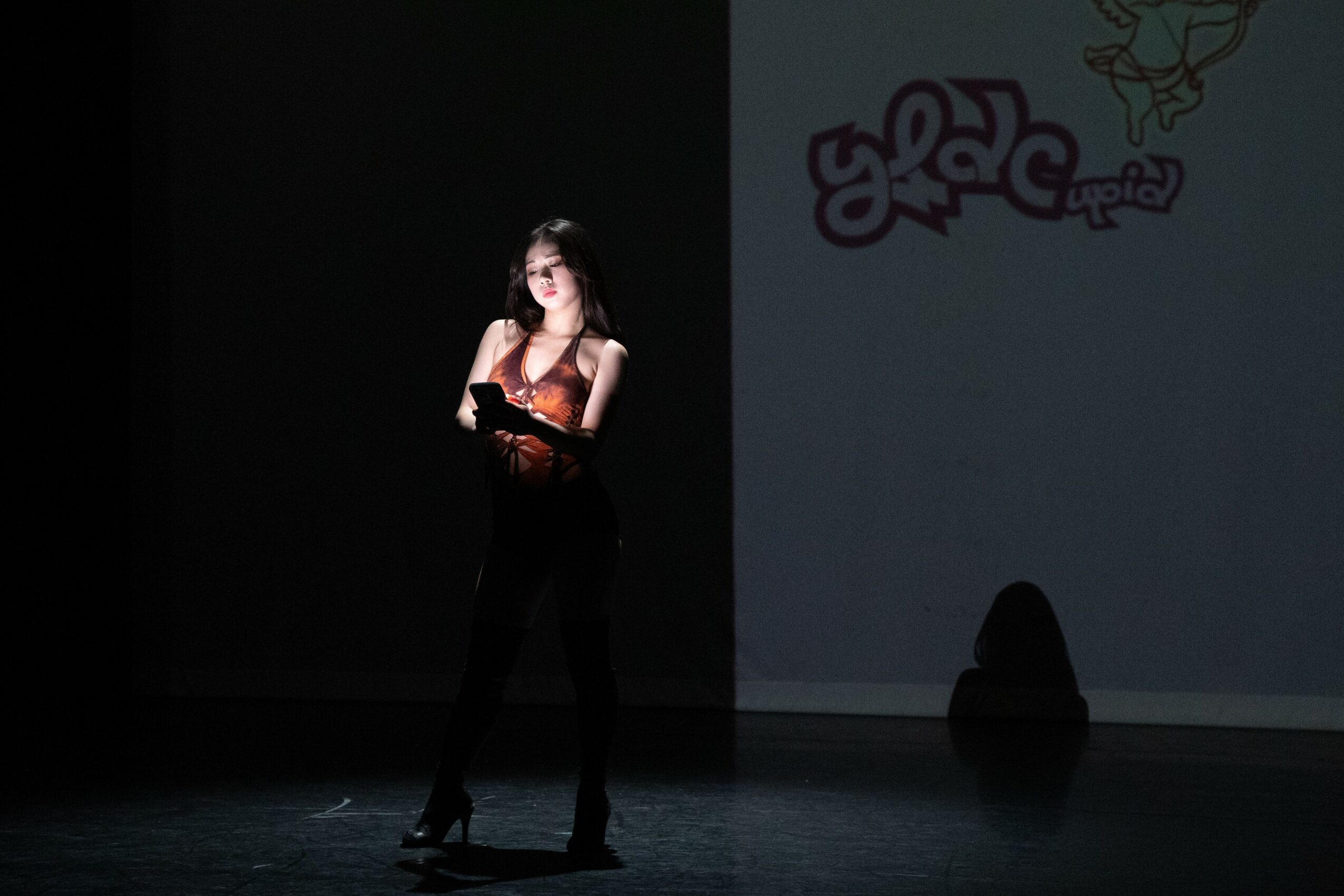 A woman stands on a stage with a dark background, illuminated by a spotlight, with graffiti on the wall behind her.