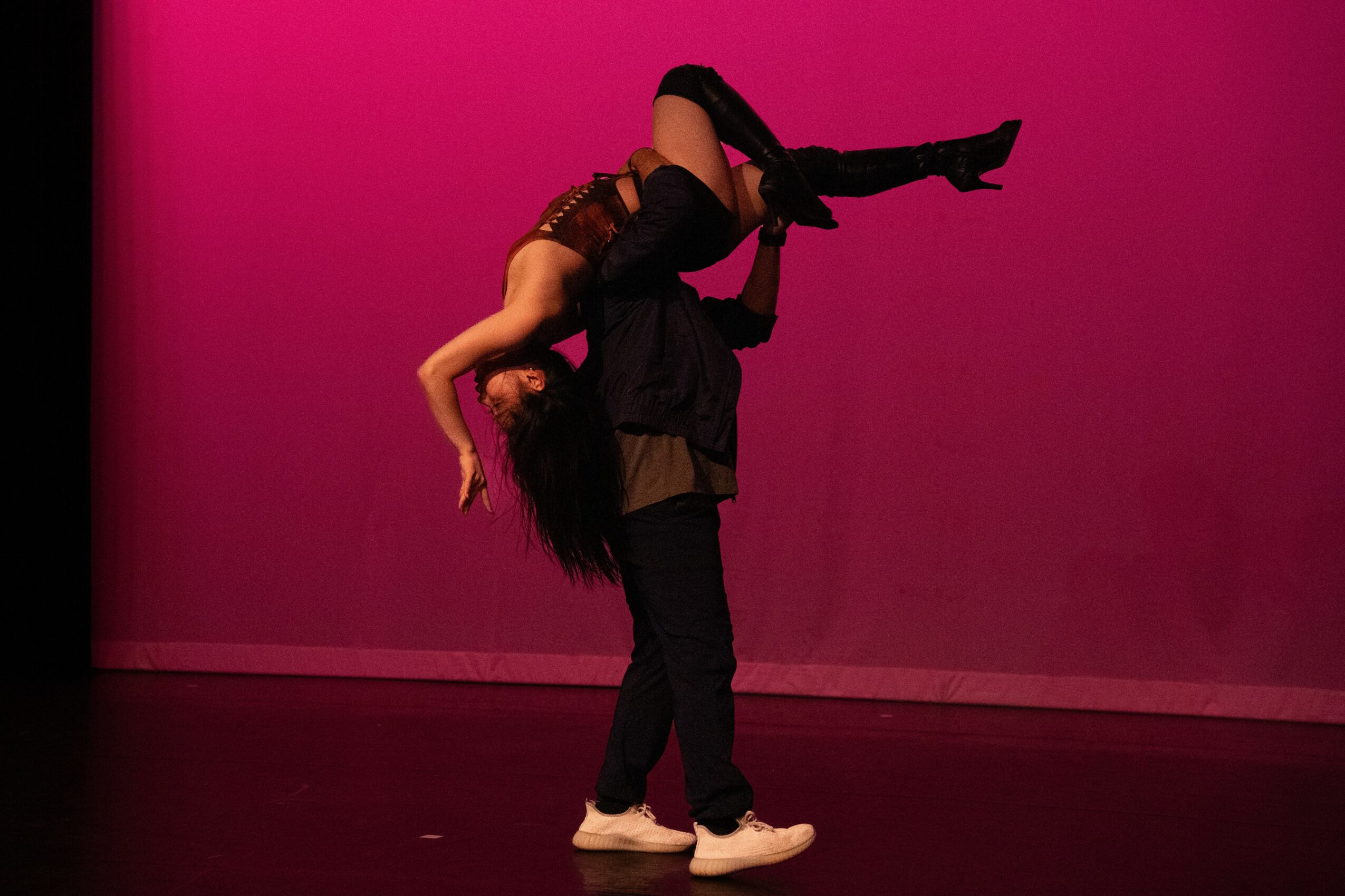 Two dancers perform on stage with a pink background, one lifting the other in an acrobatic pose.