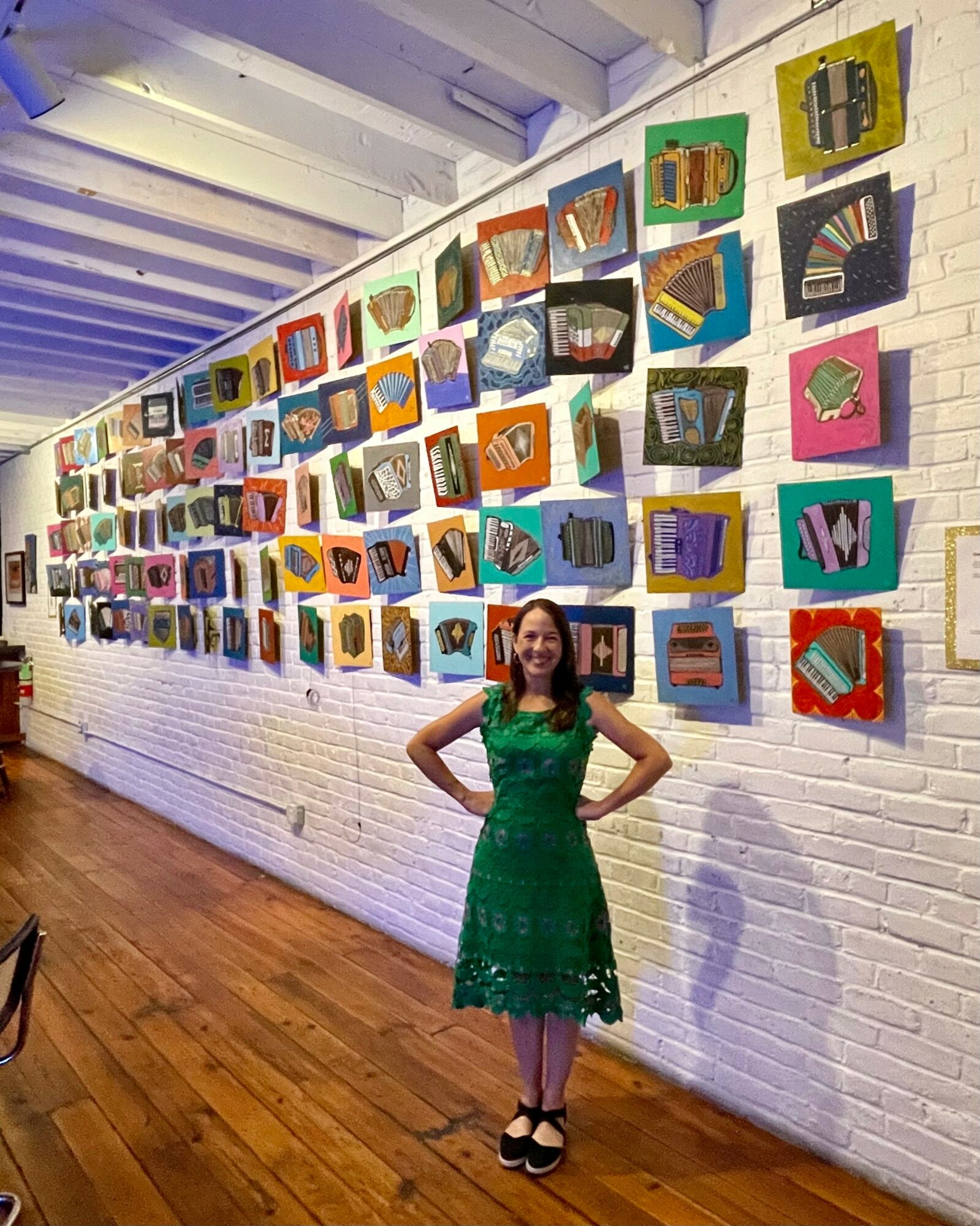 Woman in green dress stands in front of colorful artwork on a white brick wall, with wooden floor and ceiling beams visible.