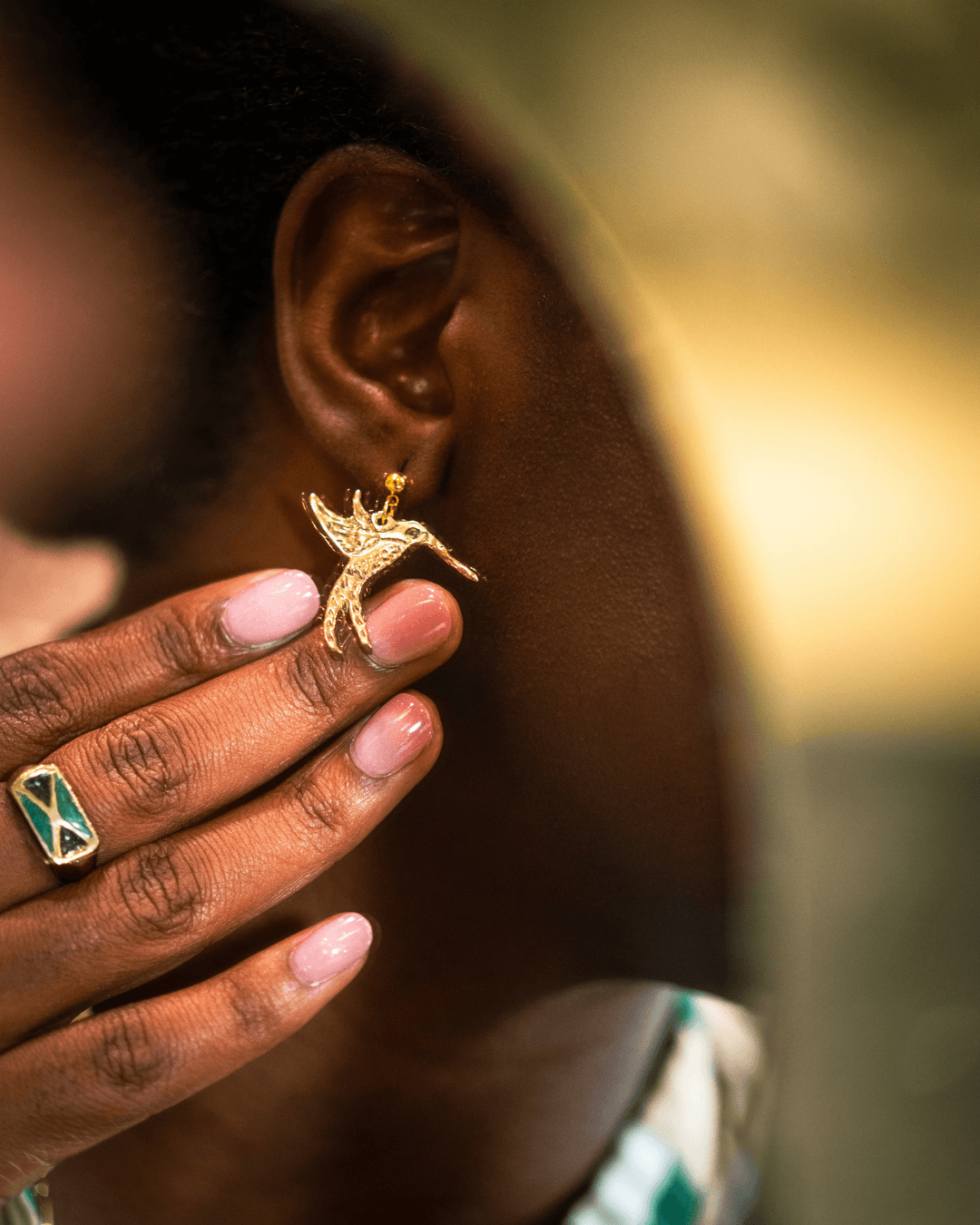 Person holding a gold deer-shaped earring near their ear, wearing a ring with a green and white design.