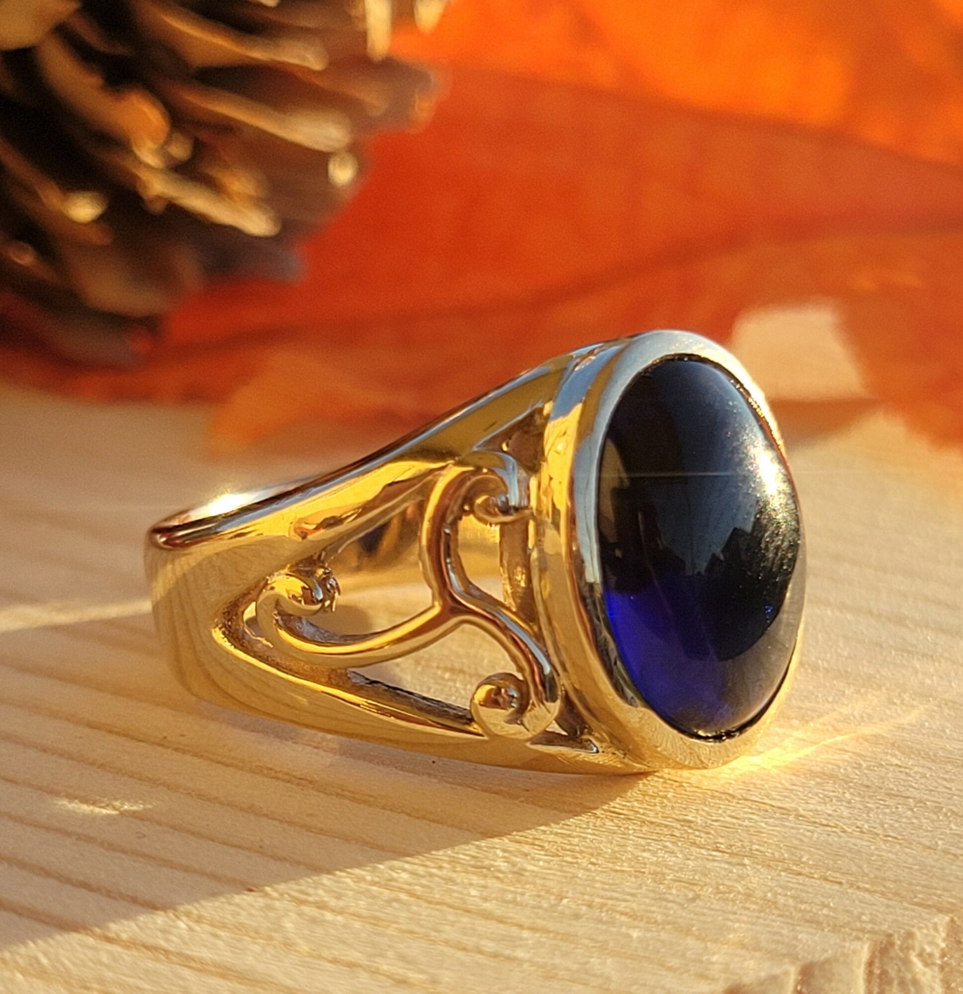 Gold ring with a large dark blue oval gemstone, resting on a wooden surface with a pine cone in the background.