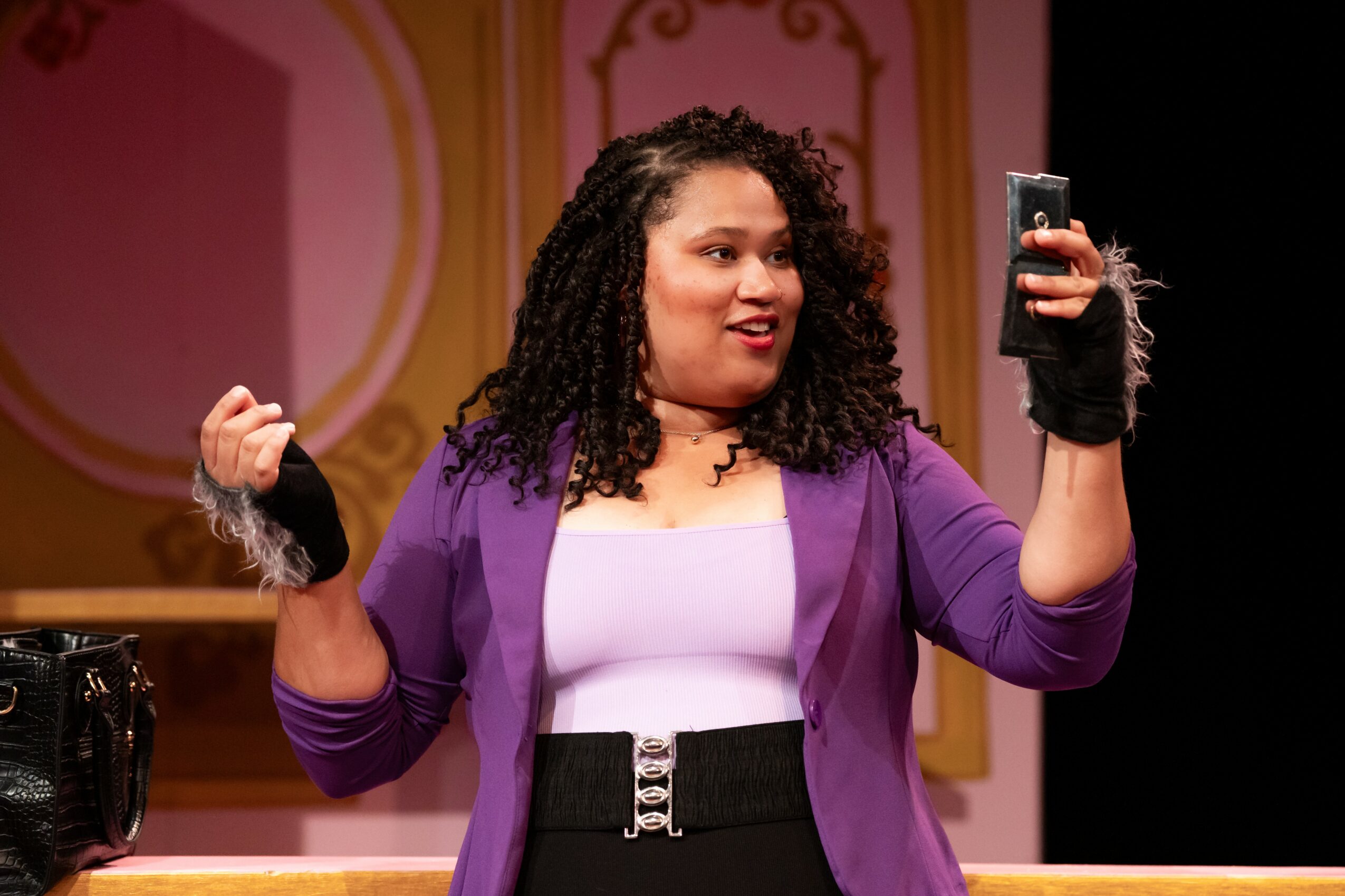 Woman with curly hair taking a selfie on stage, wearing a purple blazer and white top, with a decorative background.
