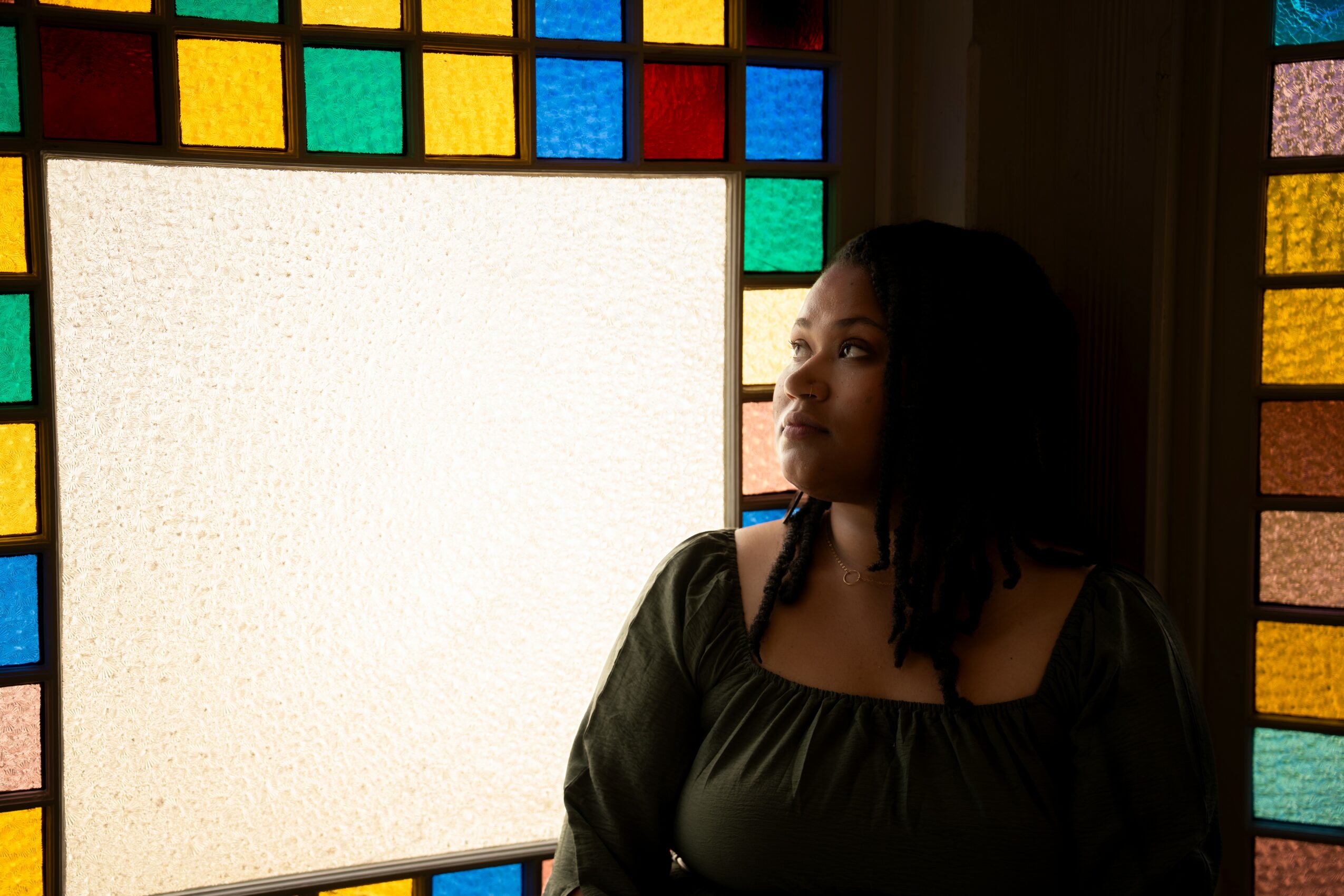 Woman with dark hair looking to the side in front of a colorful stained glass window, partially illuminated.