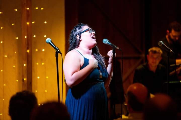 A woman singing on stage with two microphones, wearing a dark dress and glasses, with a pianist in the background.