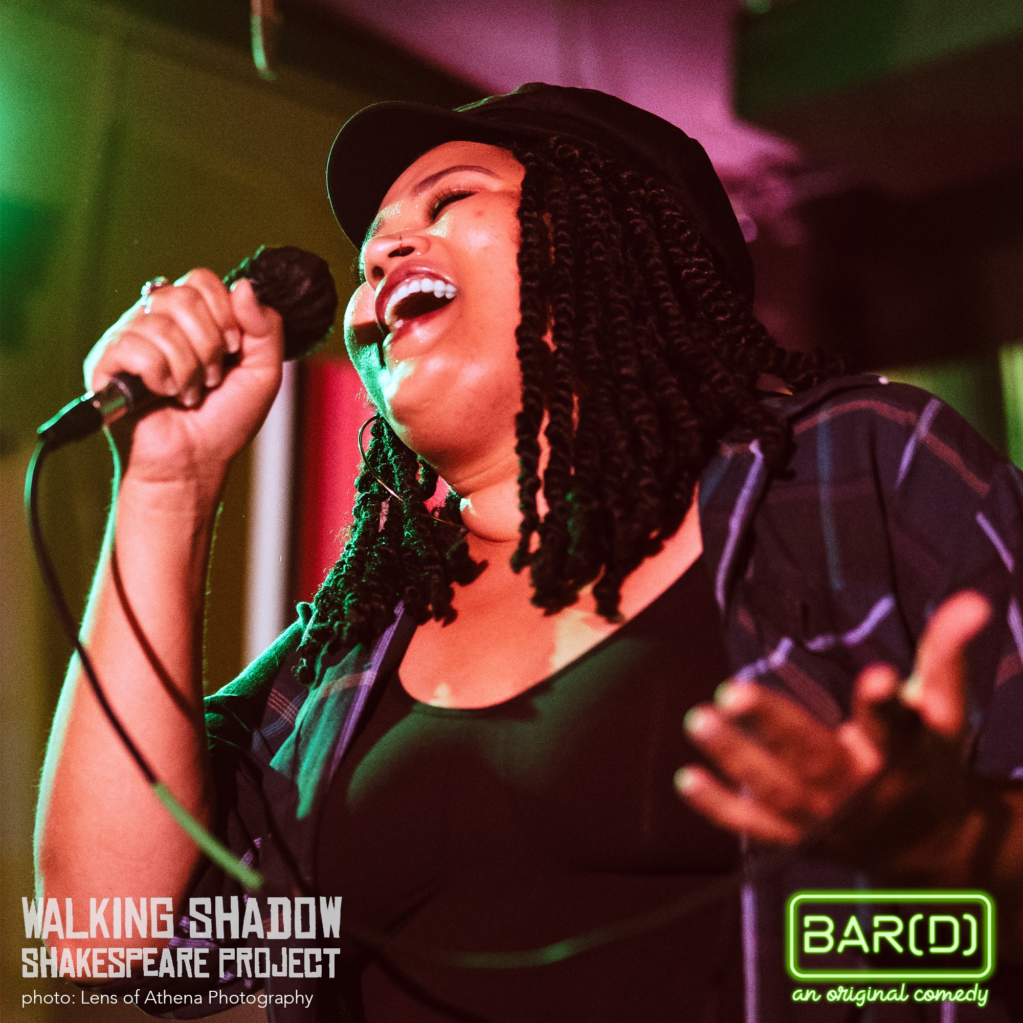 Woman singing into microphone, smiling, wearing a cap and dark clothing, colorful stage lighting in background.
