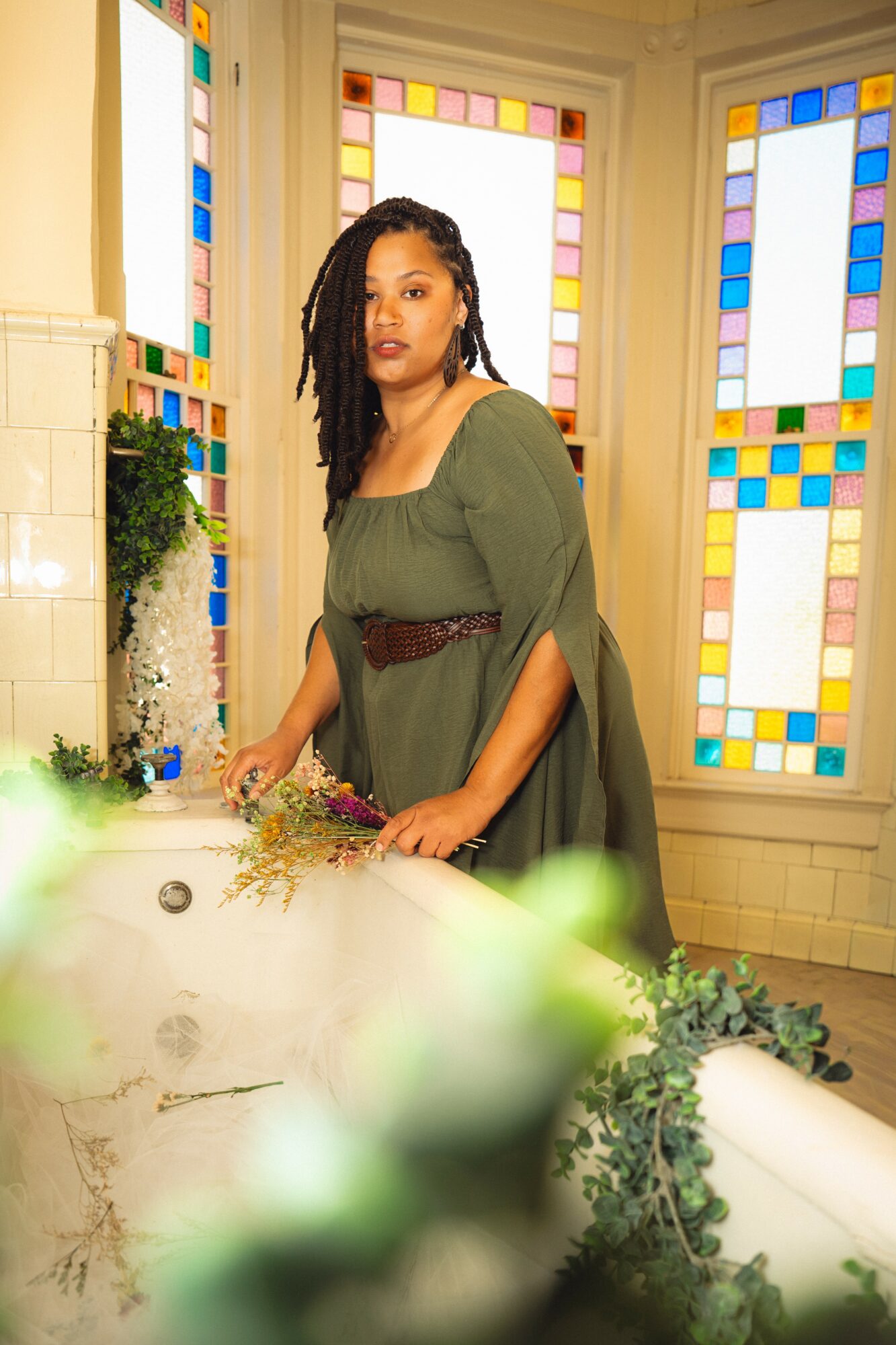 Woman with long curly hair in a green dress standing in a room with colorful stained glass windows, surrounded by plants and flowers.