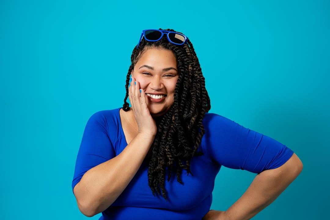 Smiling woman with long braided hair, wearing sunglasses on head and a blue shirt, against a blue background.