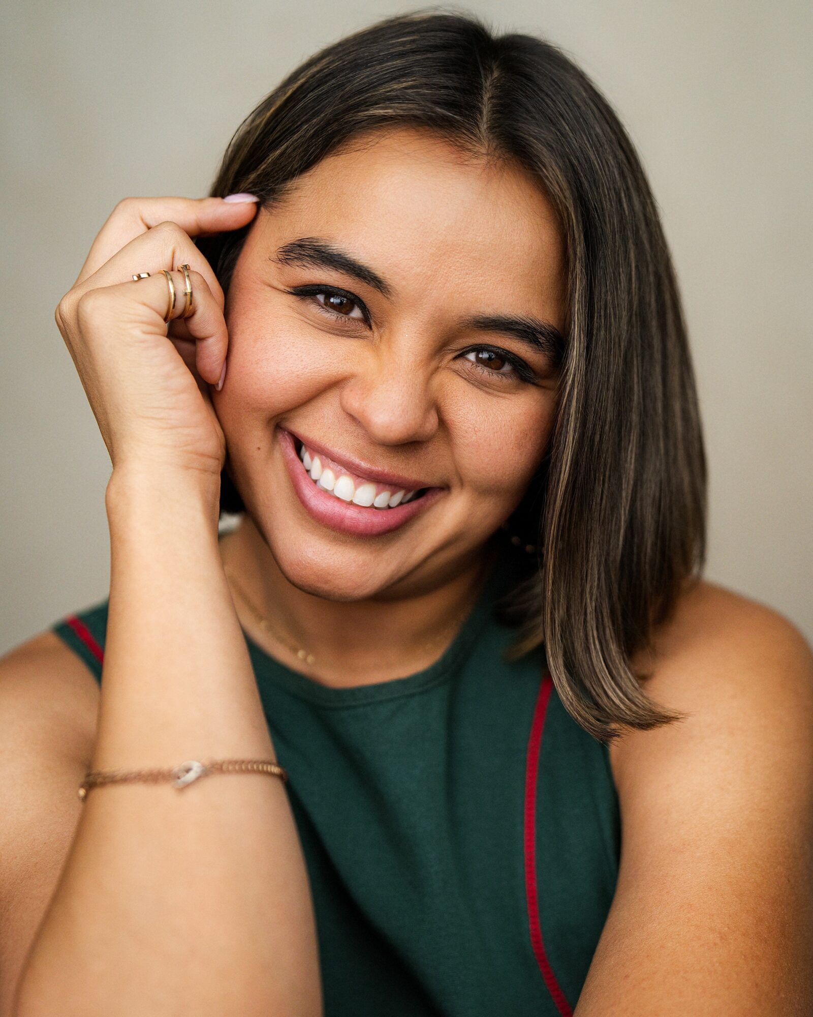 Young woman smiling, resting her head on her hand, wearing a green top, with shoulder-length dark hair.