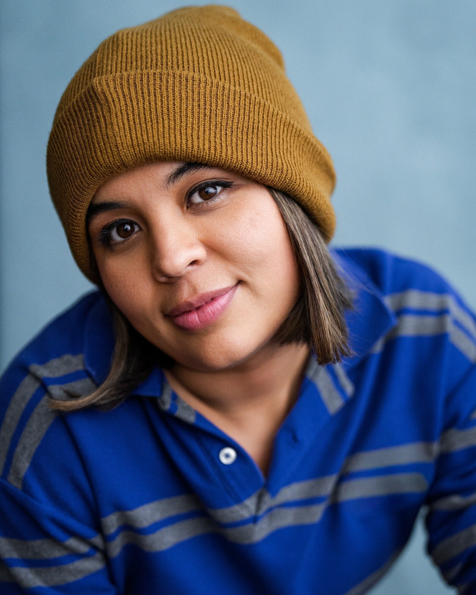 Woman wearing a mustard beanie and blue striped shirt, smiling softly, against a light blue background.