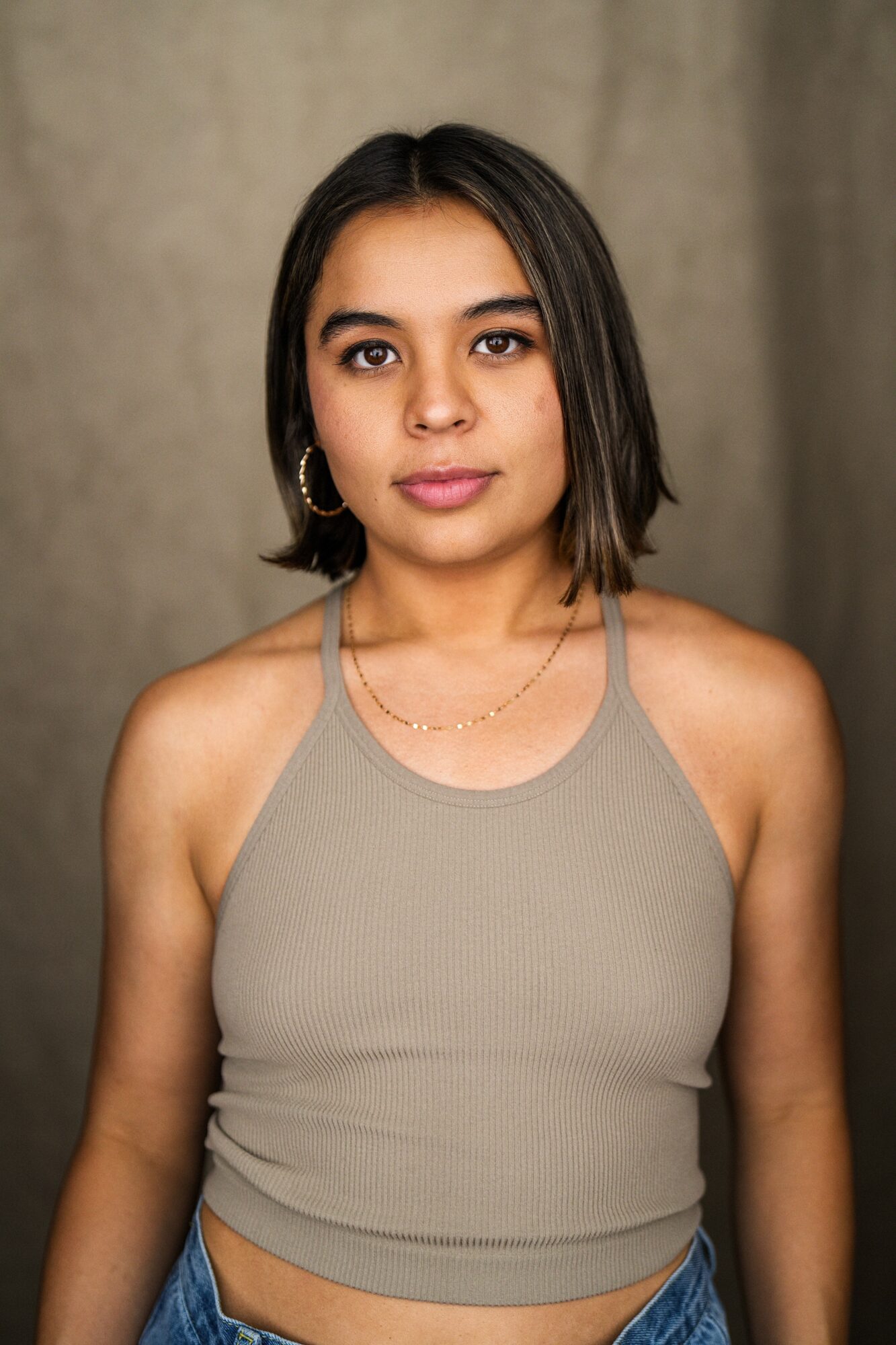 Young woman with shoulder-length dark hair, wearing a beige tank top and gold jewelry, standing against a neutral background.