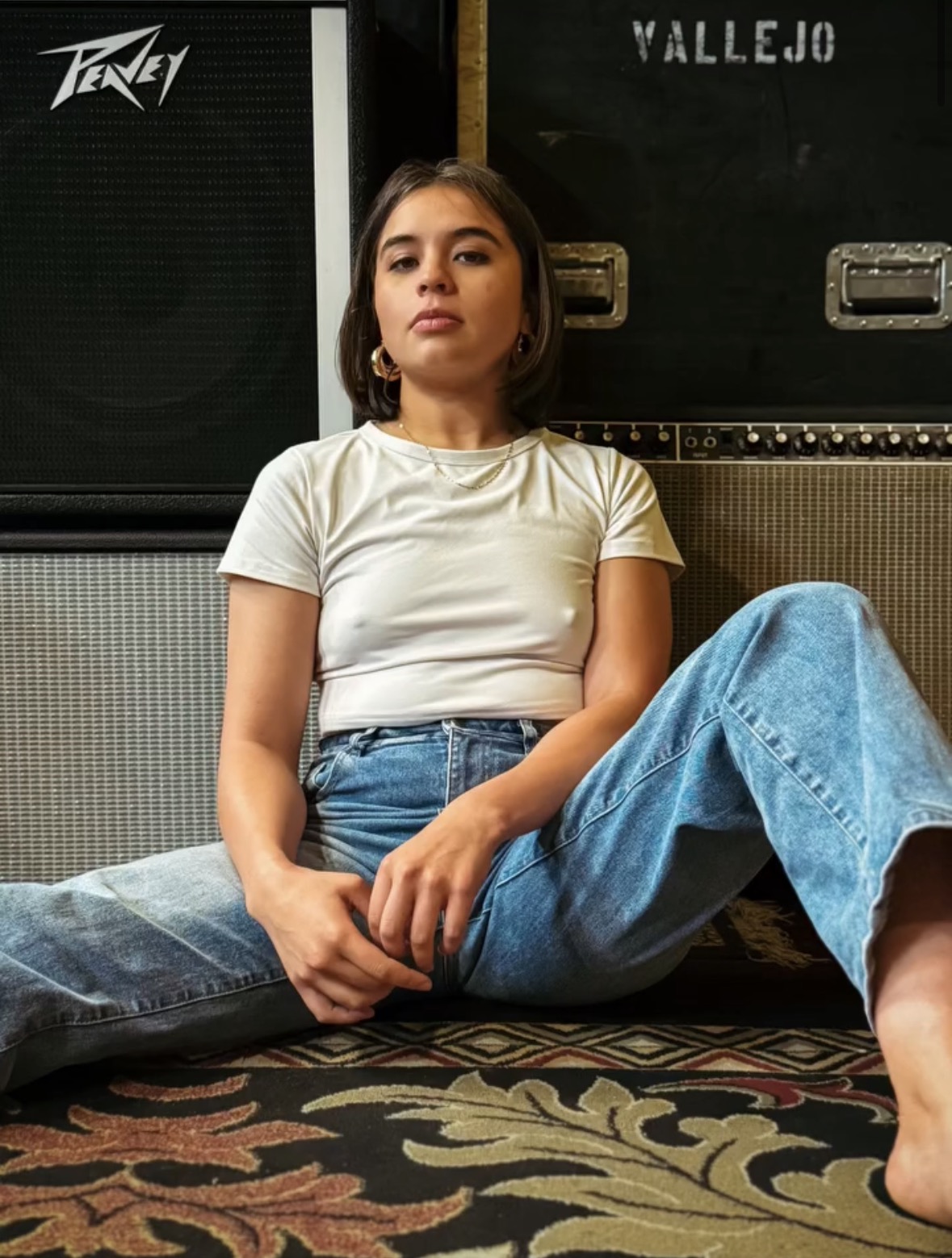 Young woman sitting on the floor with musical equipment behind her, wearing a white t-shirt and blue jeans.