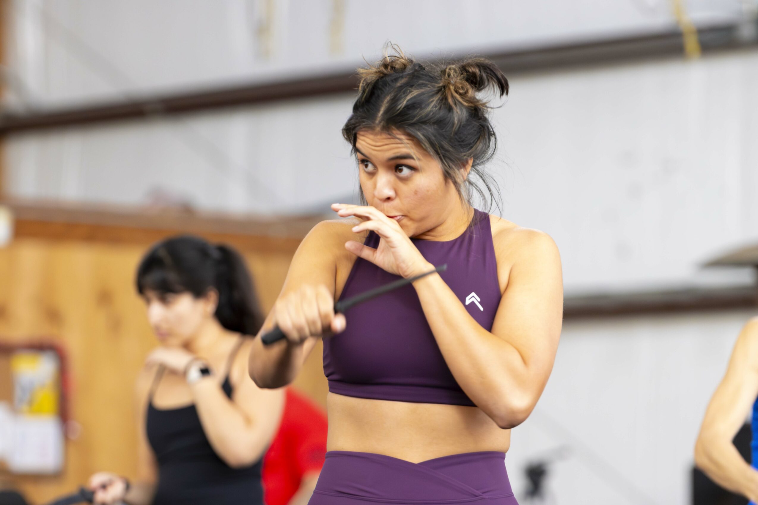 Two women in athletic clothing in a gym, one in foreground with hand near mouth, another in background stretching.
