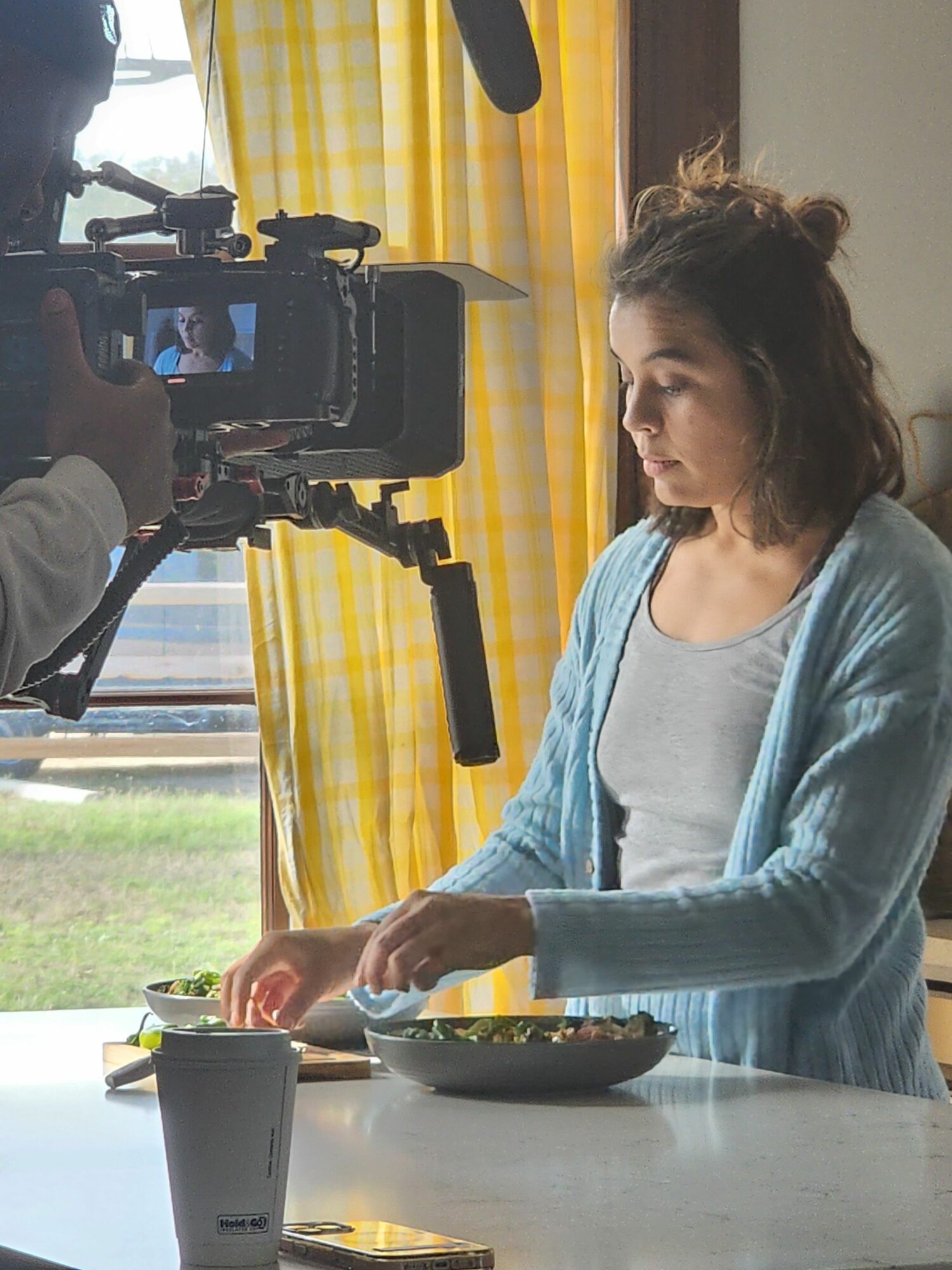 Woman with curly hair in a blue cardigan and gray shirt preparing food in a kitchen with yellow curtains.