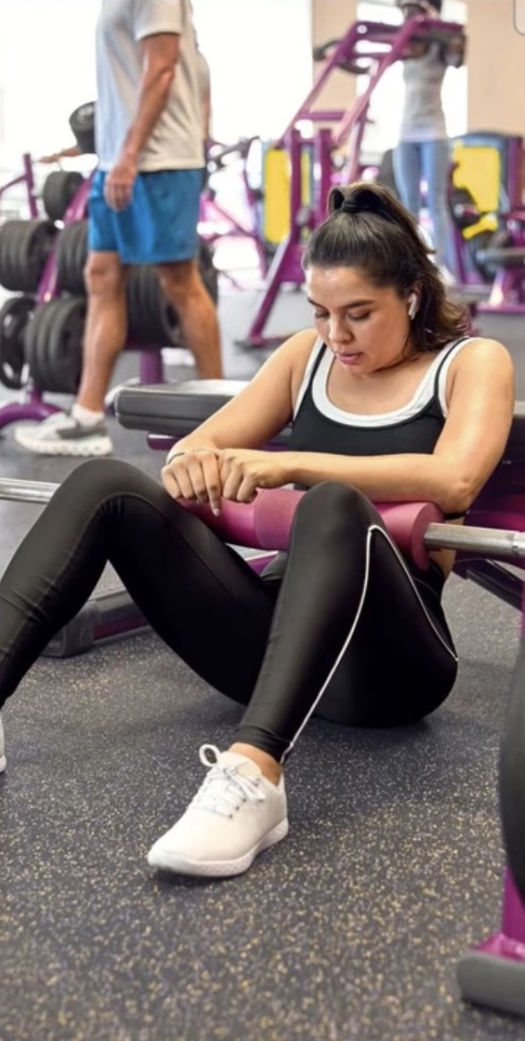Young woman sitting on the gym floor, adjusting her shoe, with gym equipment and a man in the background.