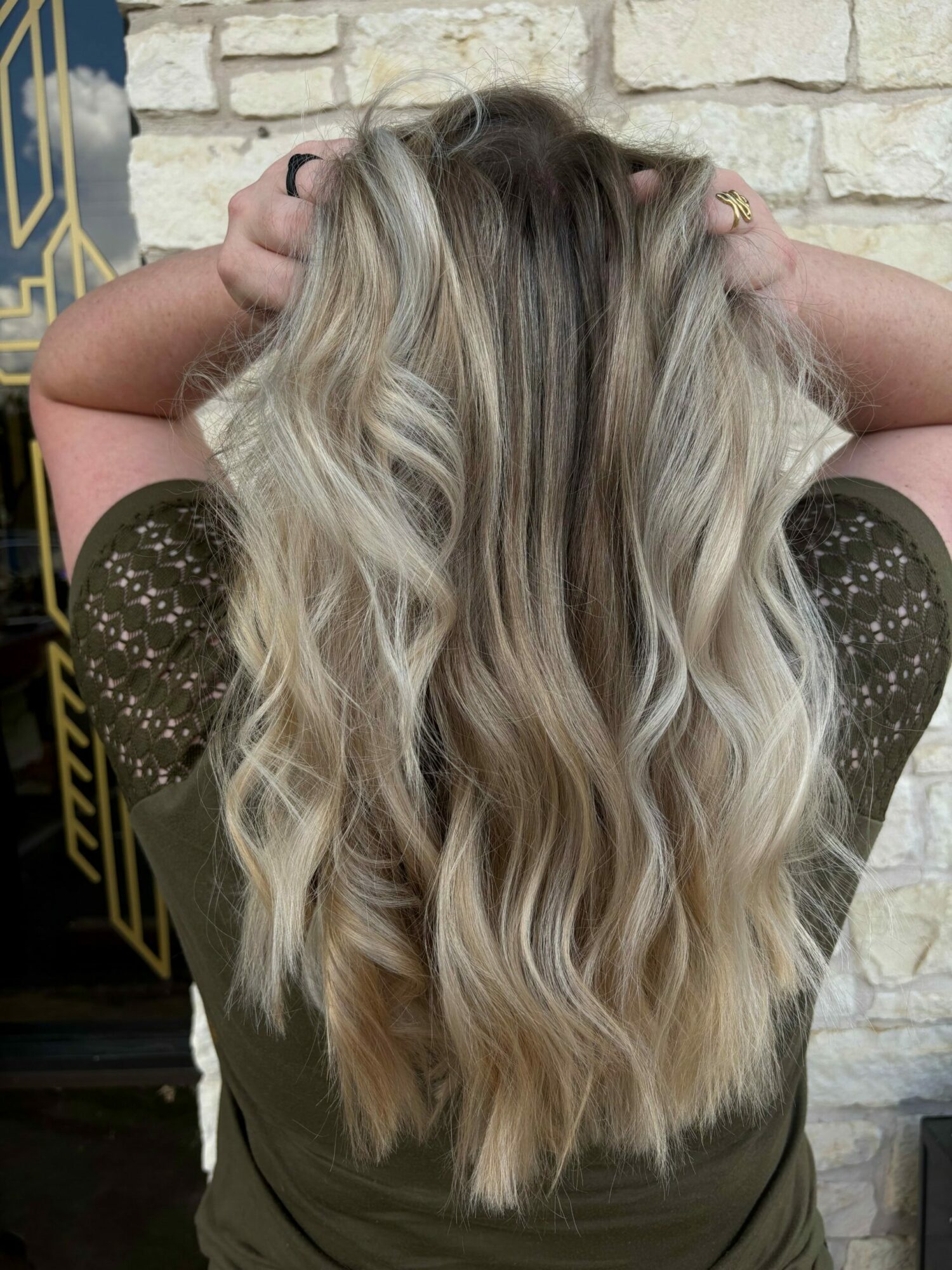 Person with long, wavy, blonde hair resting on arm, wearing a dark top with sheer sleeves, against a stone wall.
