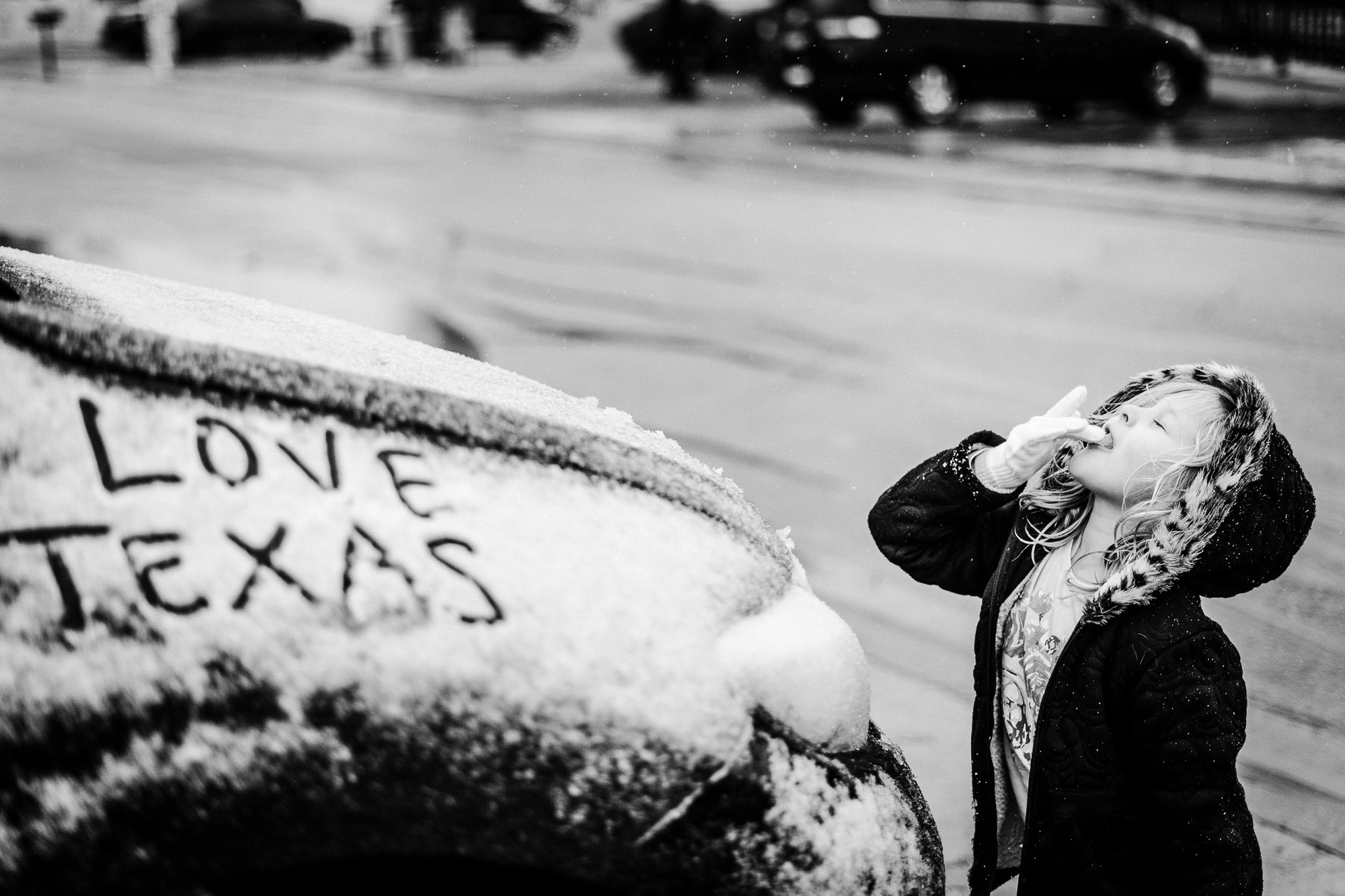 Child blowing snow off a car with 'LOVE TEXAS' written in snow, on a street with moving cars in background.