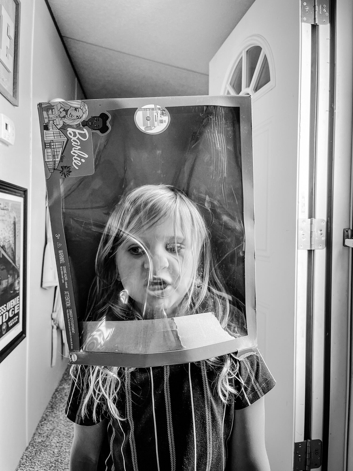 Child with long hair wearing a striped shirt looks through a plastic window on a box, with a surprised expression.