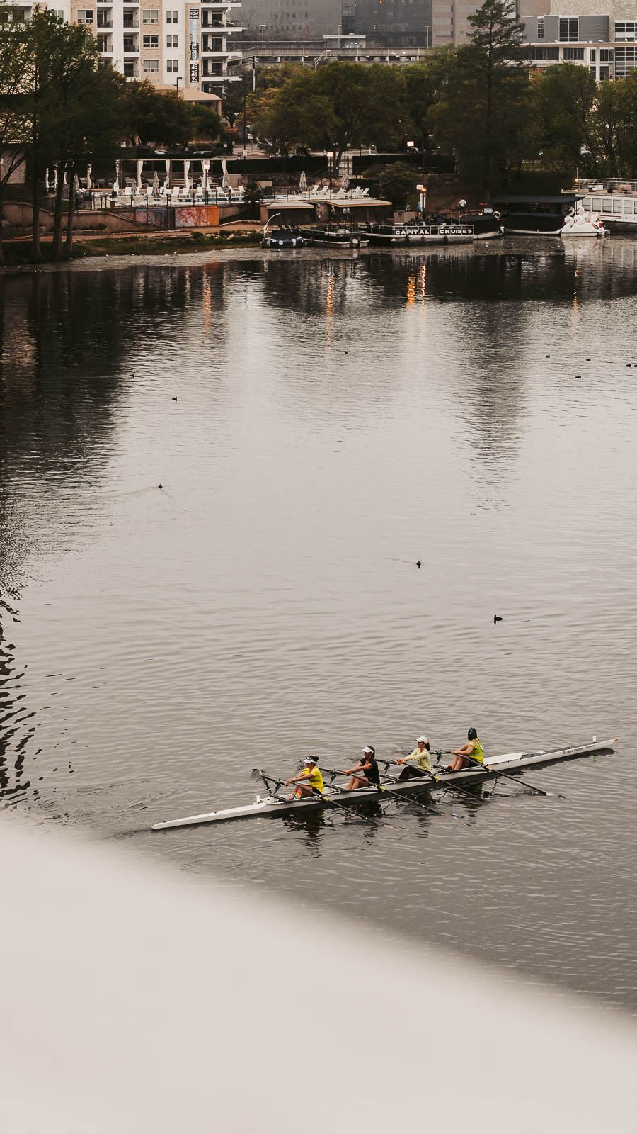 Four people rowing a boat on a river with buildings and trees in the background.