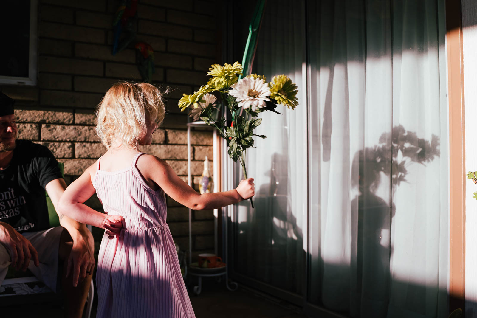 Child with blonde hair in a pink dress reaches for a bouquet of flowers on a window ledge, shadow cast on curtain.