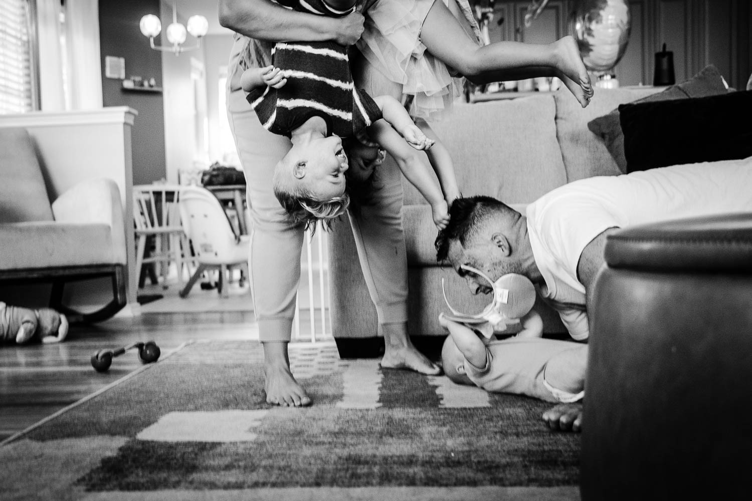 Child standing upside down with head on the floor, another person kneeling nearby, and a child holding a balloon in a living room.