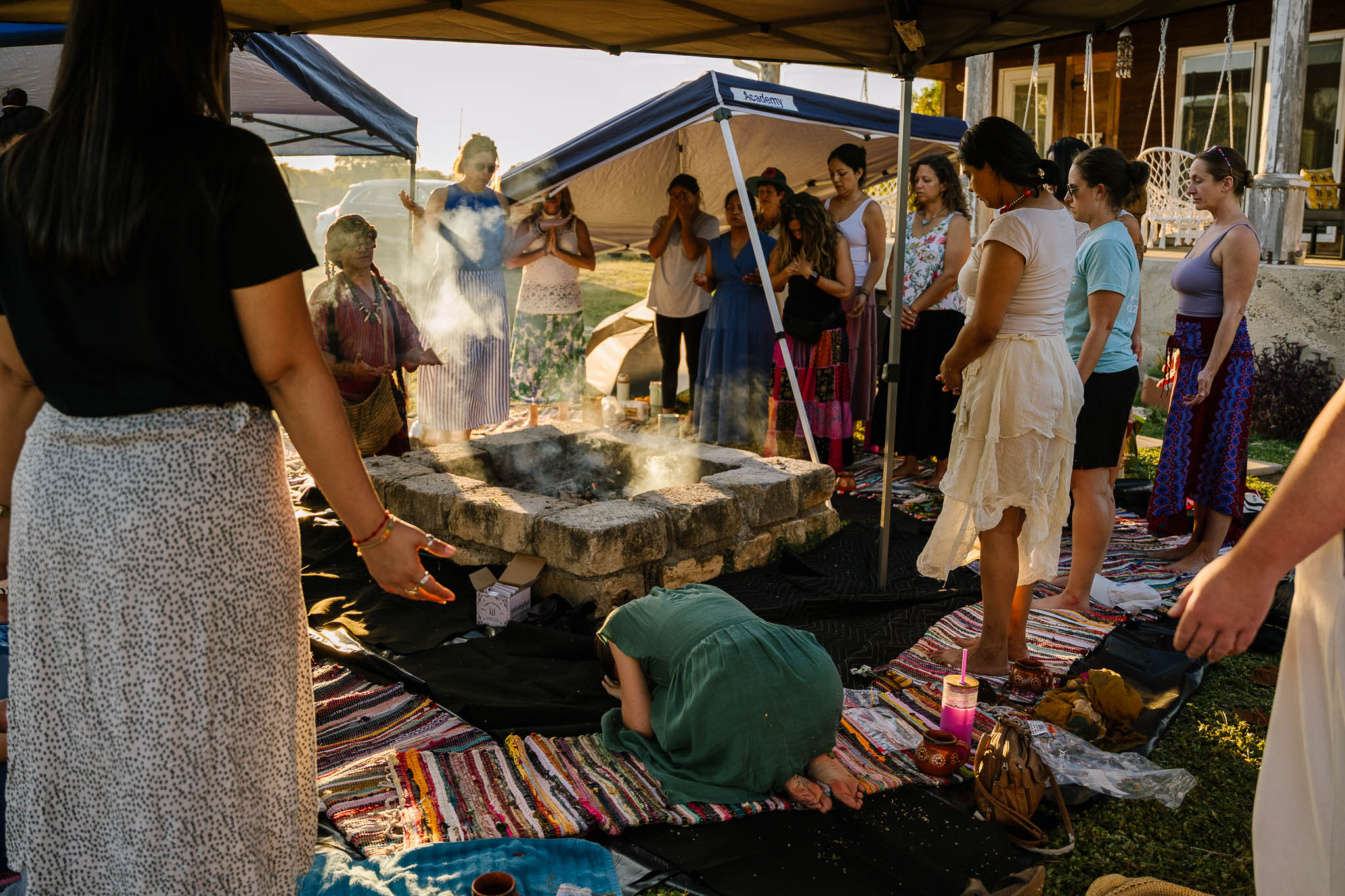 Group of people gathered outdoors around a stone fire pit, some standing and some sitting on colorful rugs, with tents in background.