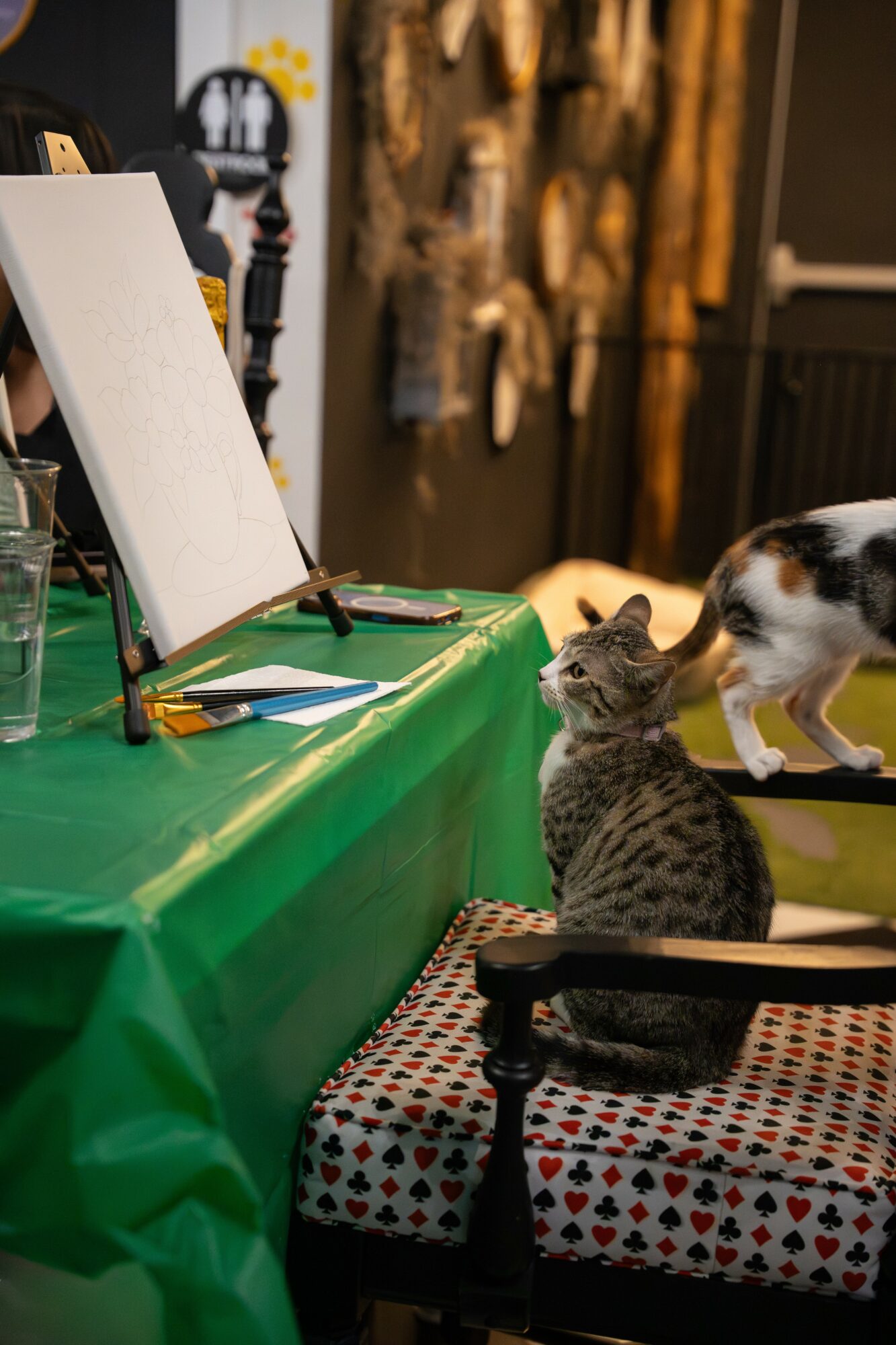 Two cats sitting on a chair with a green tablecloth, in front of an easel and art supplies, in a room with wooden wall decor.
