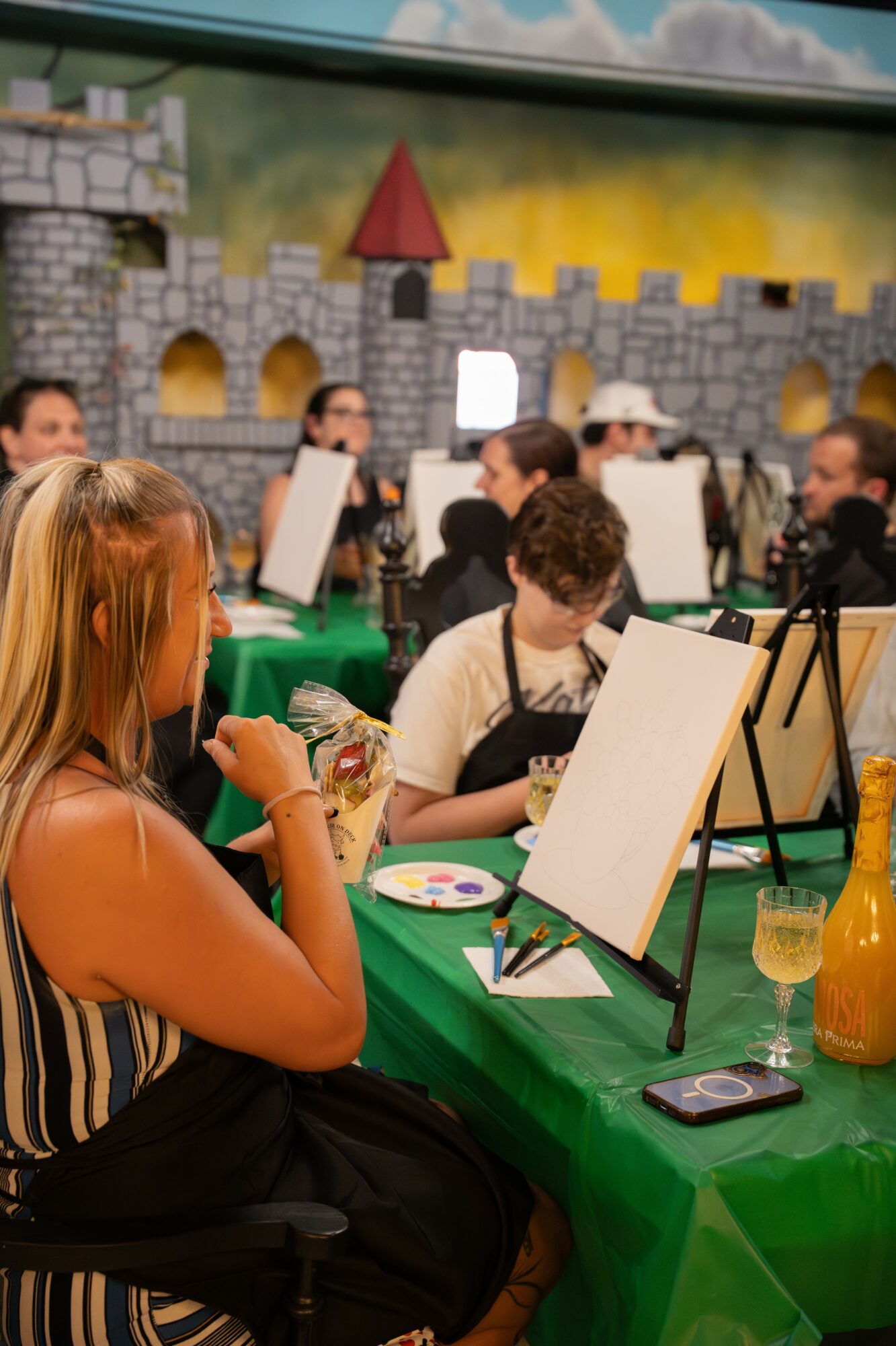 People painting on canvases at a table with a castle mural background, some wearing hats, and drinks on the table.