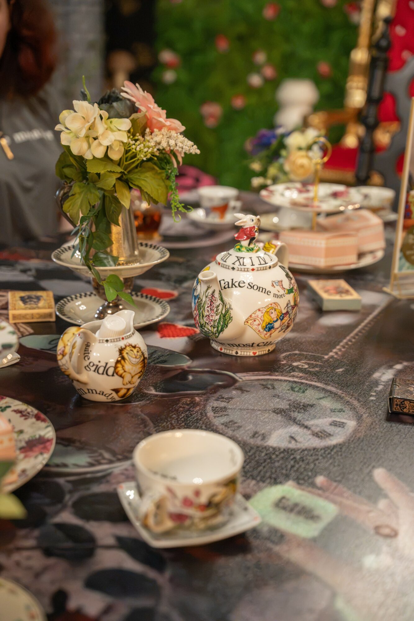 Decorative teapots and a floral bouquet on a table with various dishes and utensils, outdoors with greenery in the background.