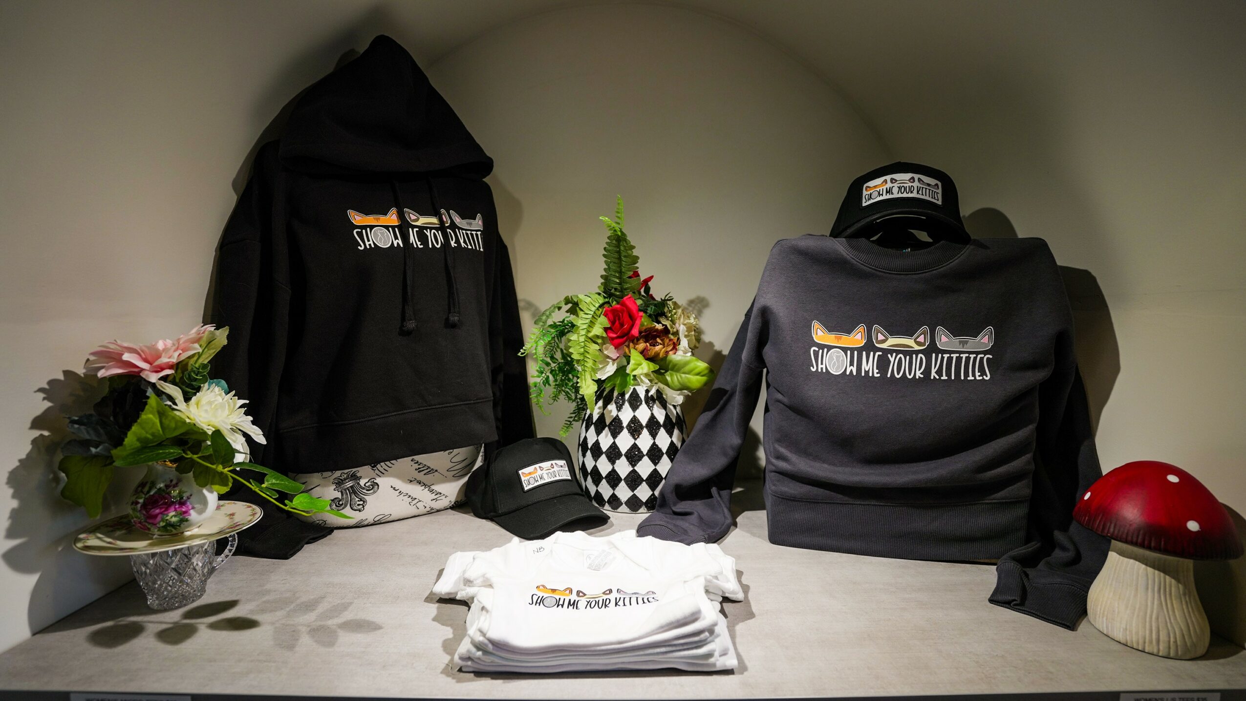 Display with black hoodie, backpack, flowers, cap, and a red object on a table.