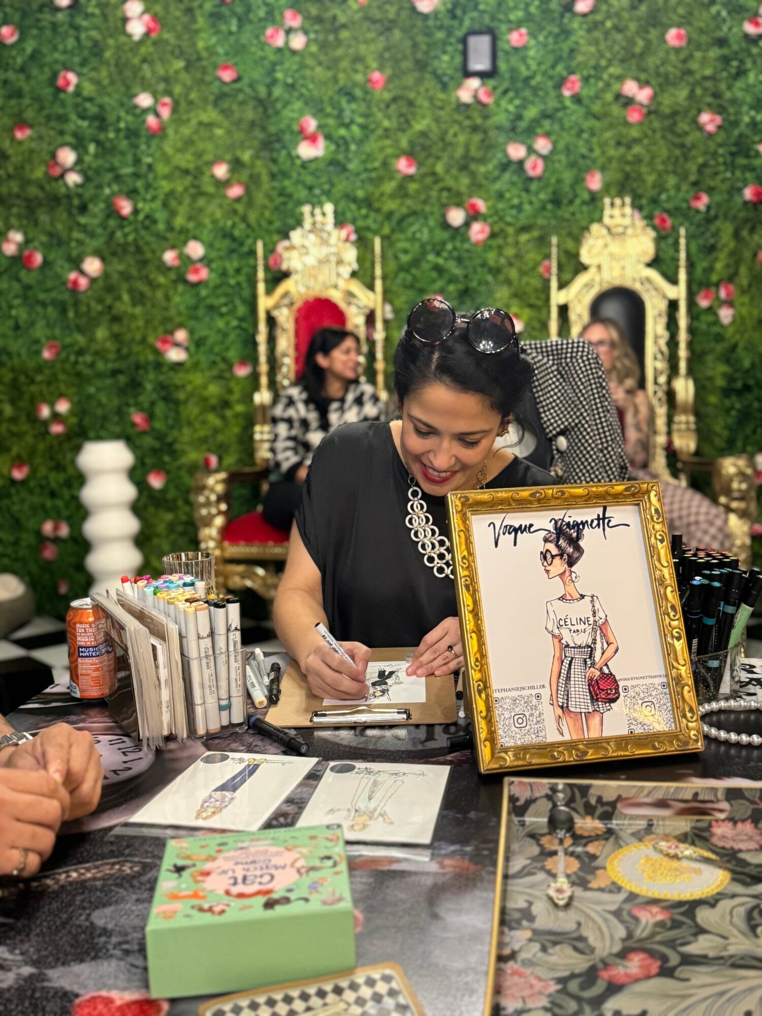 Woman signing a framed illustration at a table with a green floral backdrop and pink flowers, two women seated behind her, decorative chairs, and various items on the table.