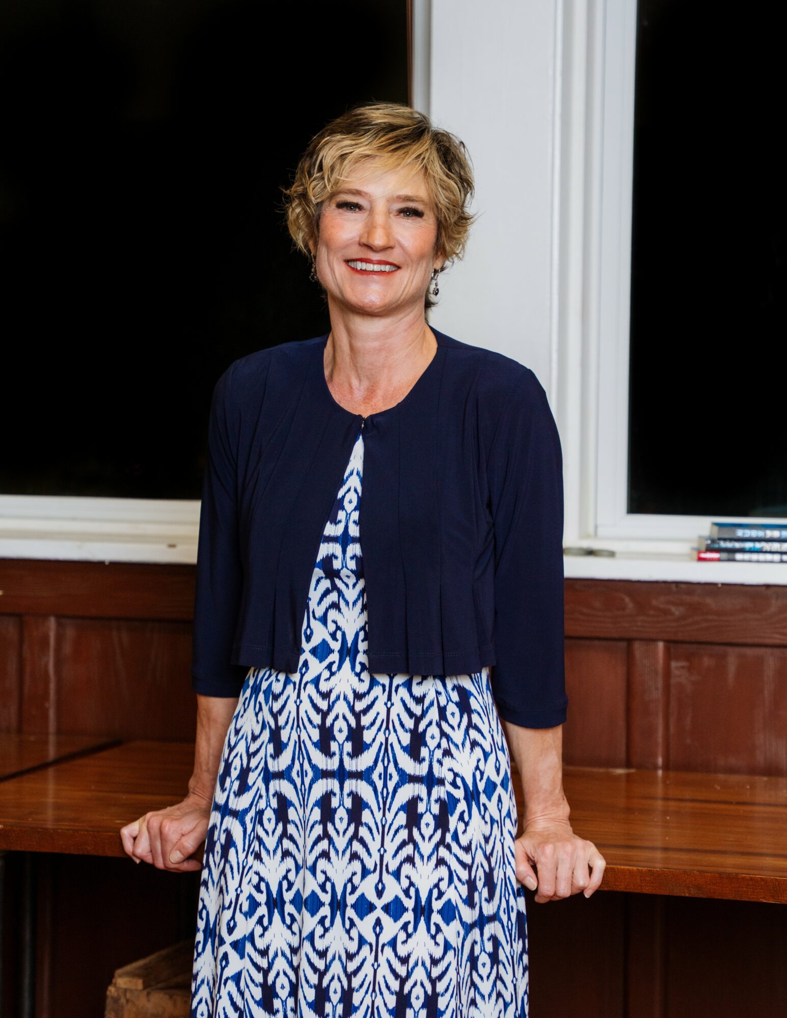 Smiling woman with short blonde hair wearing a blue and white patterned dress and navy cardigan, standing indoors.