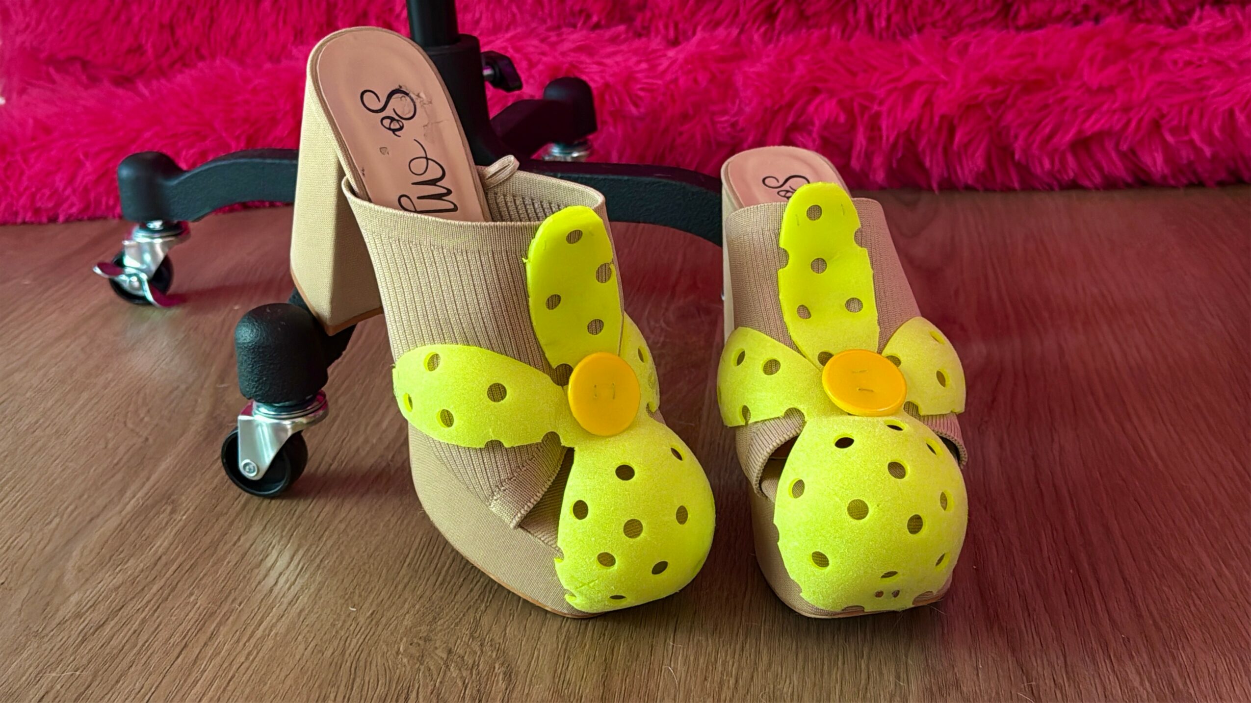 Pair of children's shoes with yellow polka-dotted bows and beige straps on a wooden surface, pink fuzzy background.