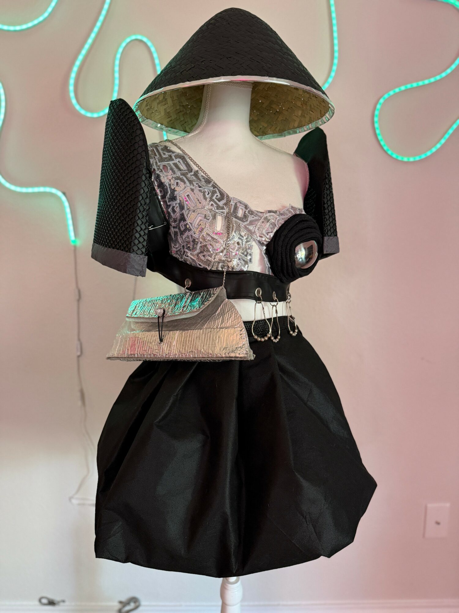 Mannequin dressed in a large black hat, patterned top, black skirt, and accessories, against a pink wall with neon lights.