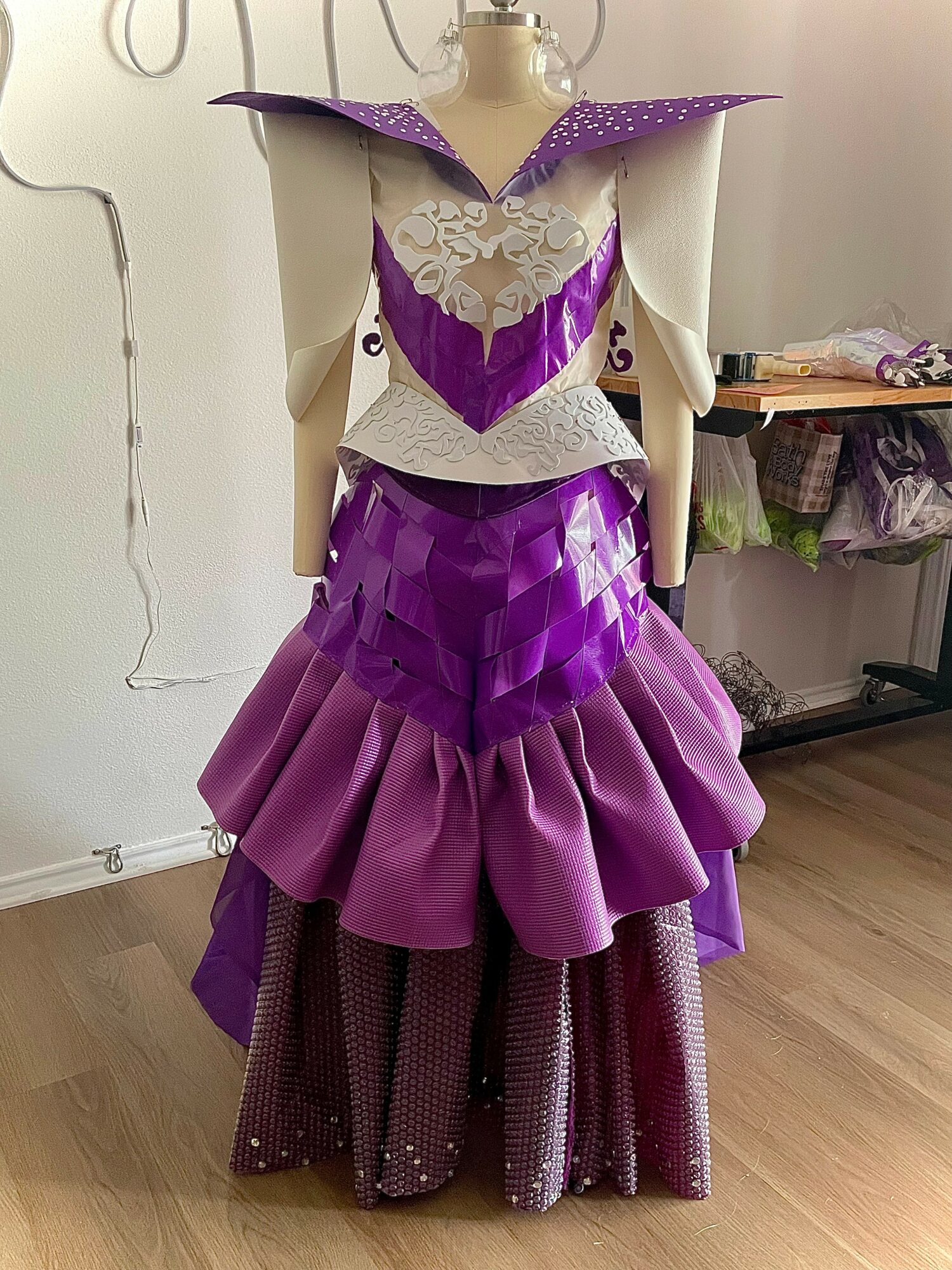 Mannequin dressed in a purple and white layered, ruffled gown with a high collar and decorative details, standing indoors.