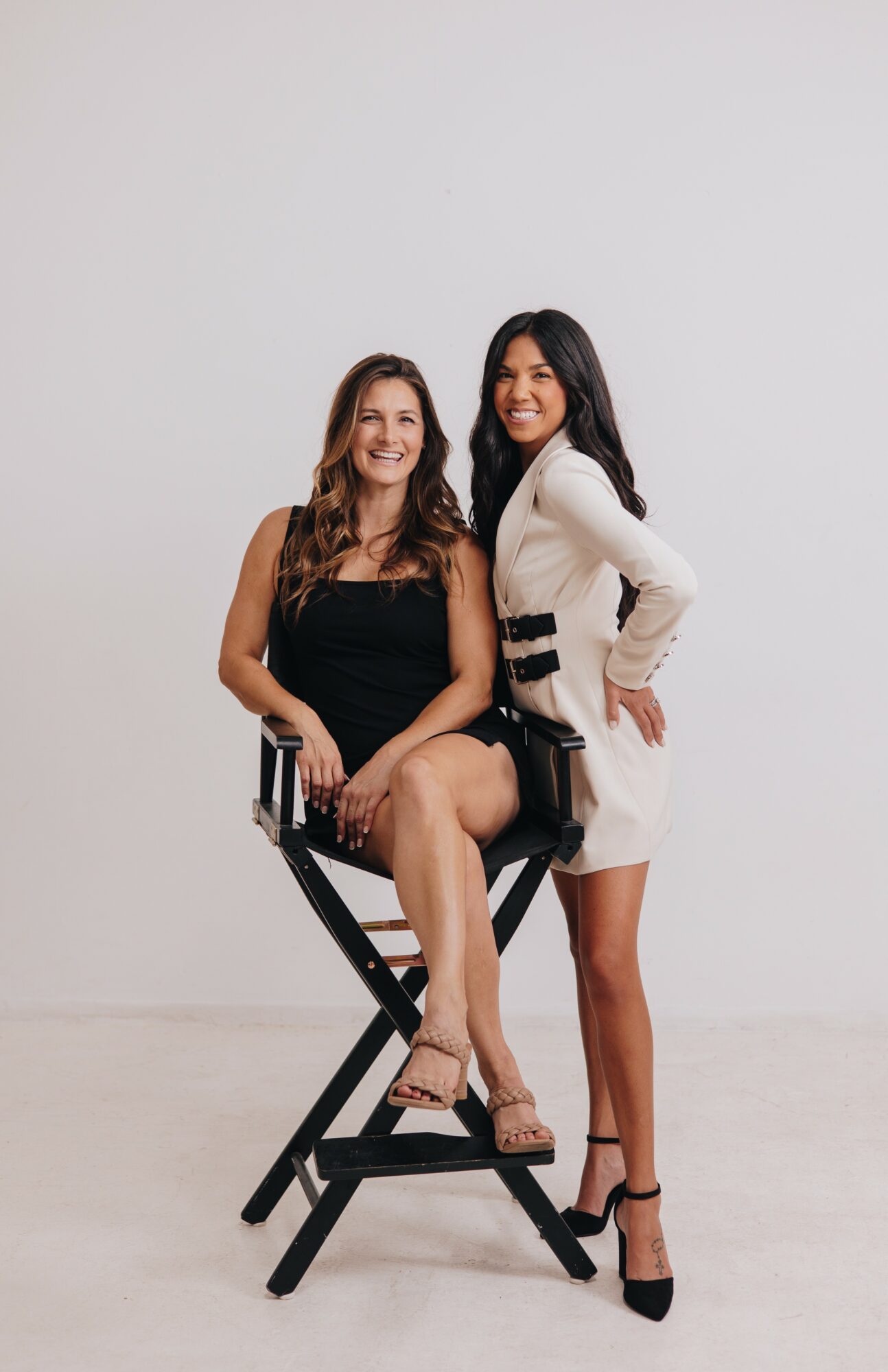 Two women pose together, one sitting on a black chair and the other standing beside her, smiling.