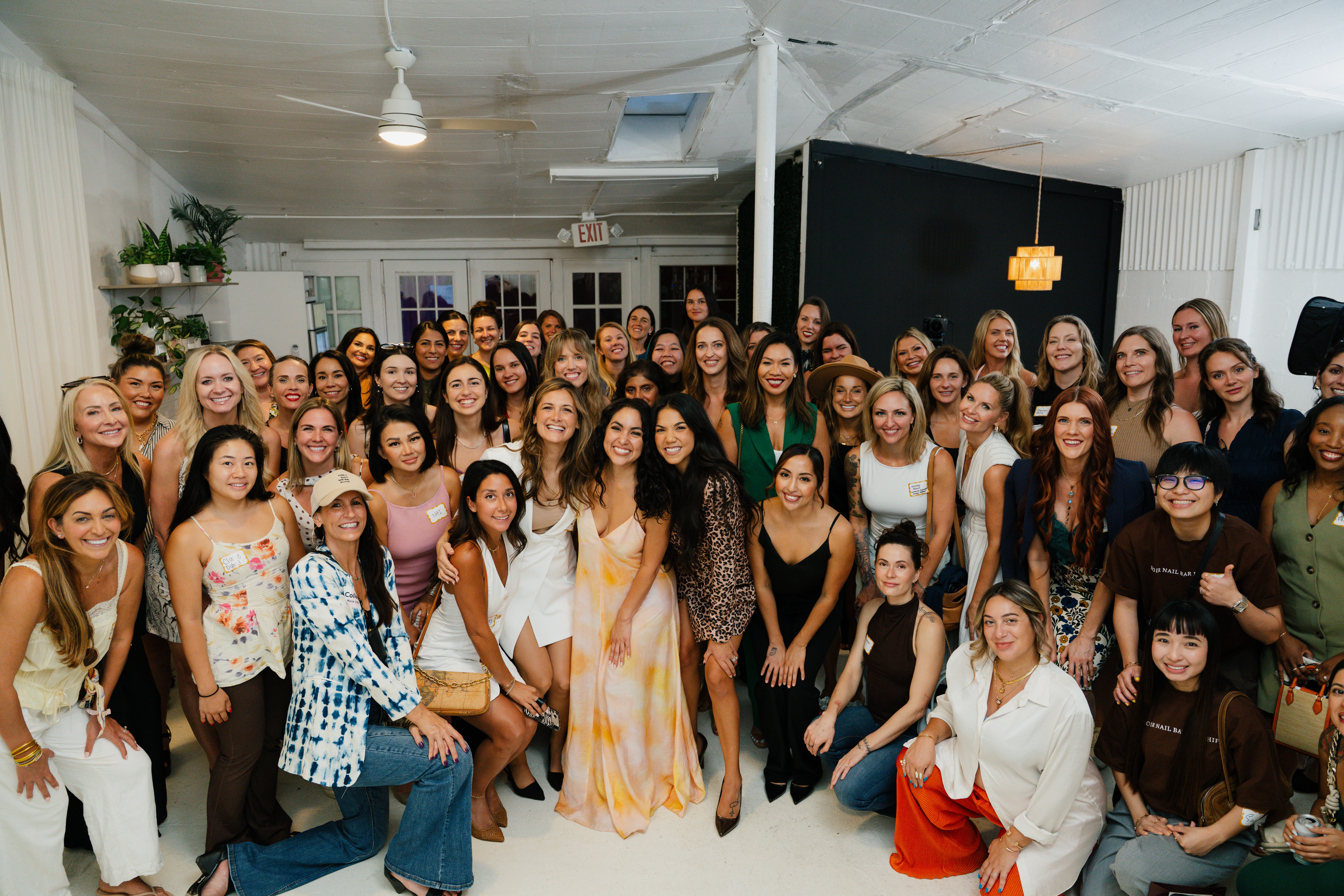 Large group of women and girls gathered indoors, smiling for a group photo, with white walls and ceiling lights.