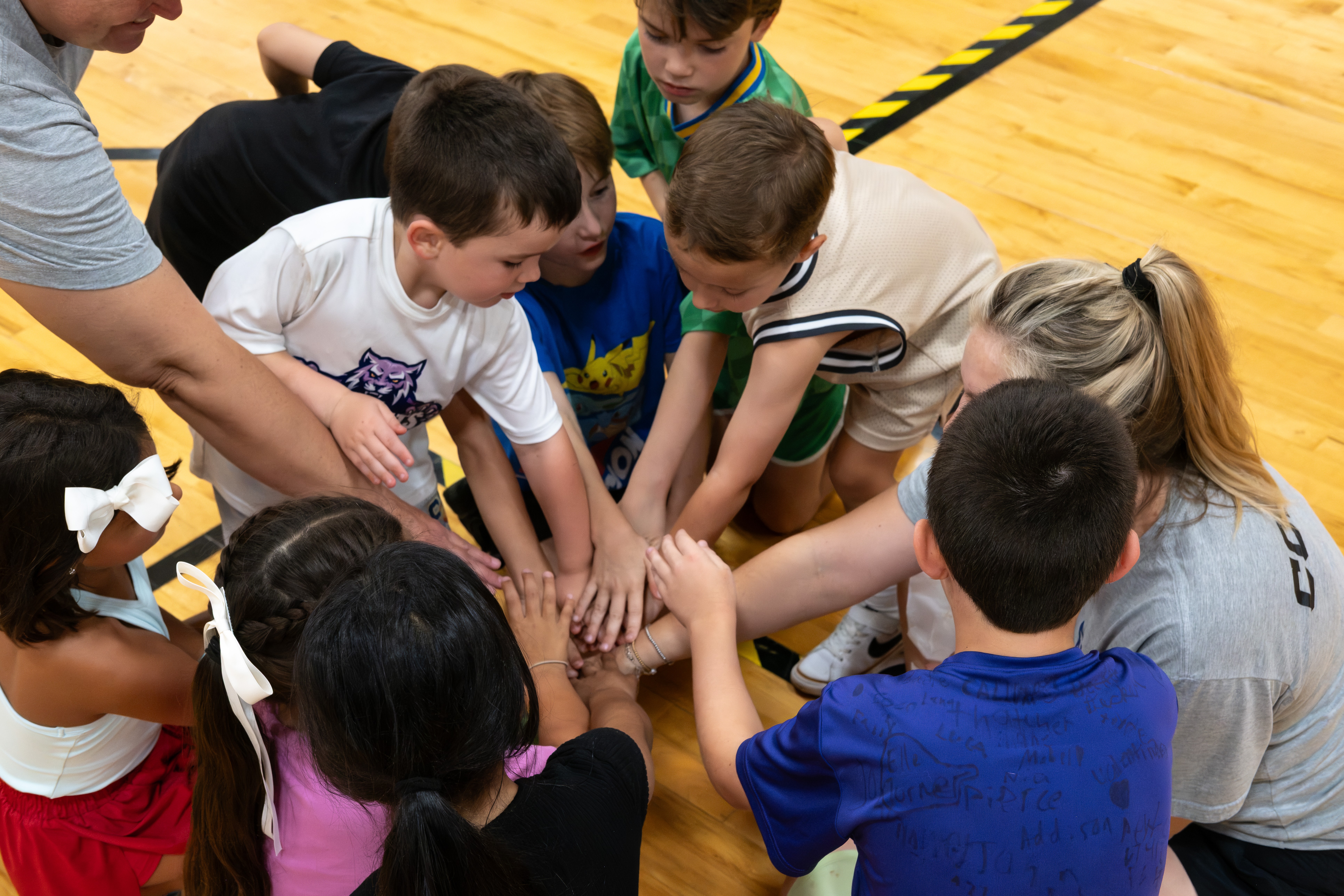 Group of children and adults with hands stacked together in a gymnasium, engaging in a team activity.