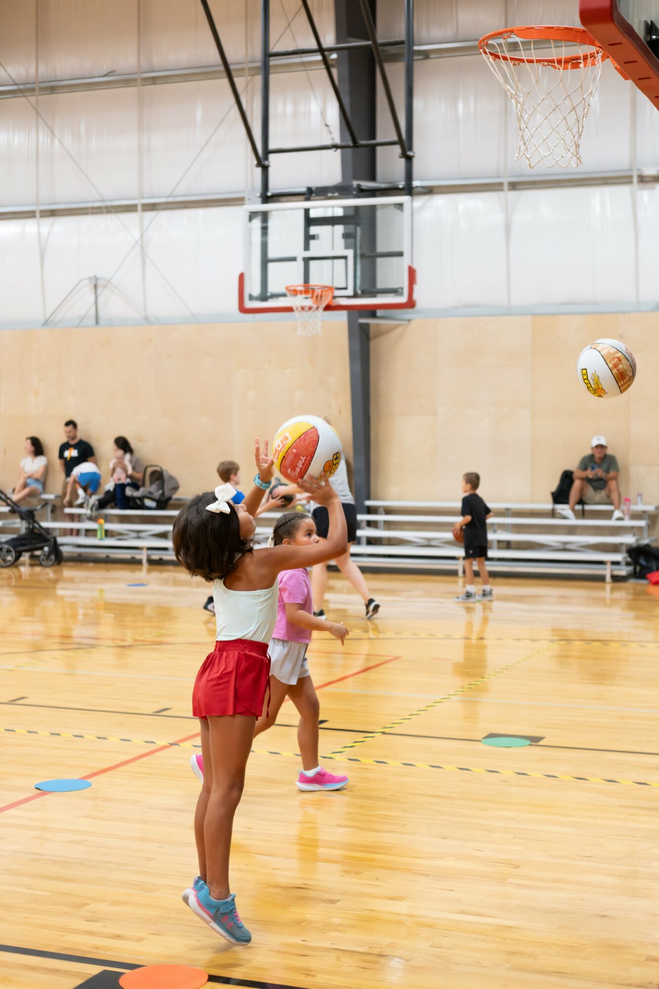 Children playing basketball in an indoor gymnasium, some jumping to shoot or catch the ball, with spectators in the background.