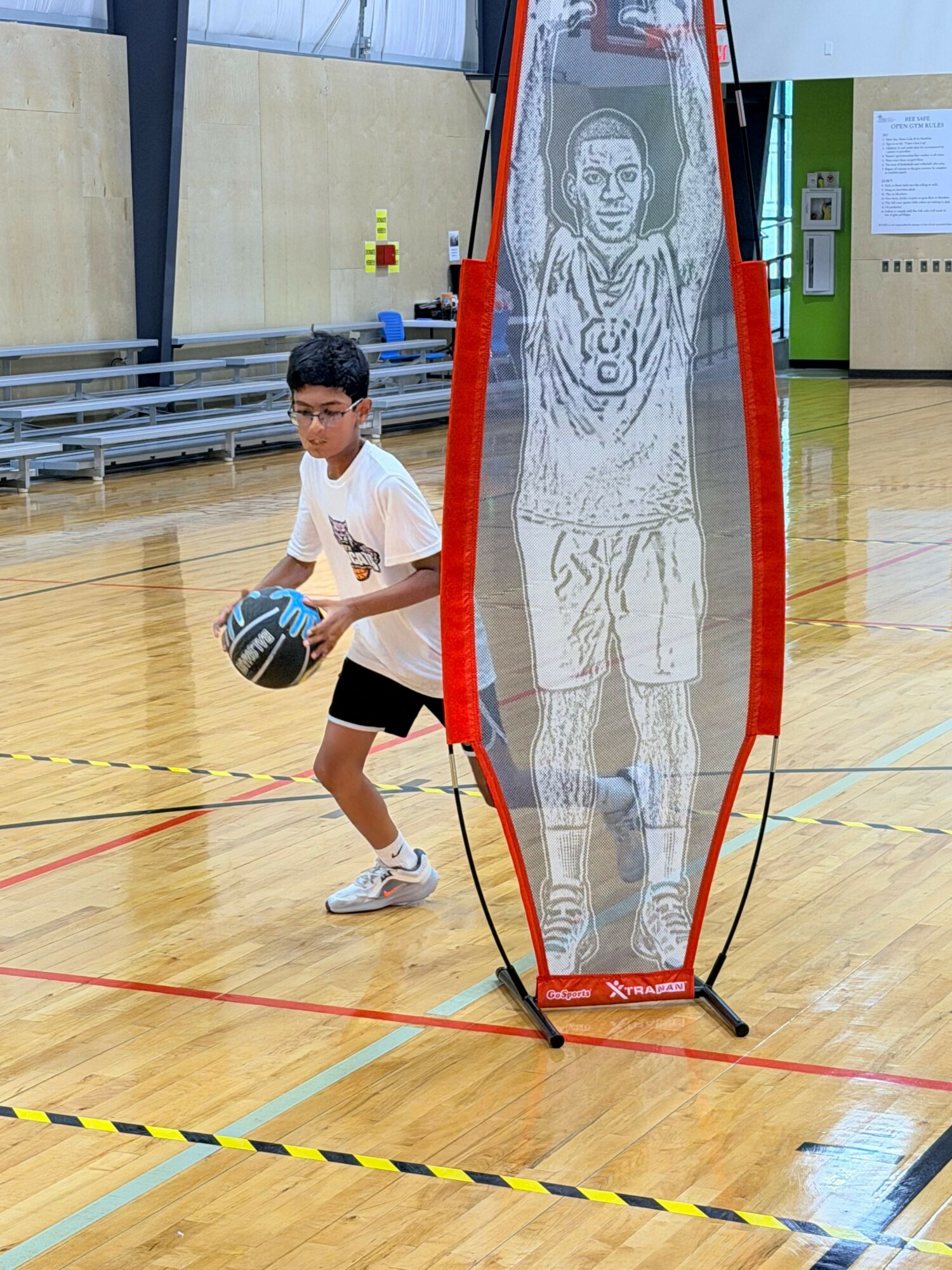 Person practicing basketball in a gym, holding a basketball, with a large banner or poster behind them, gym floor visible.