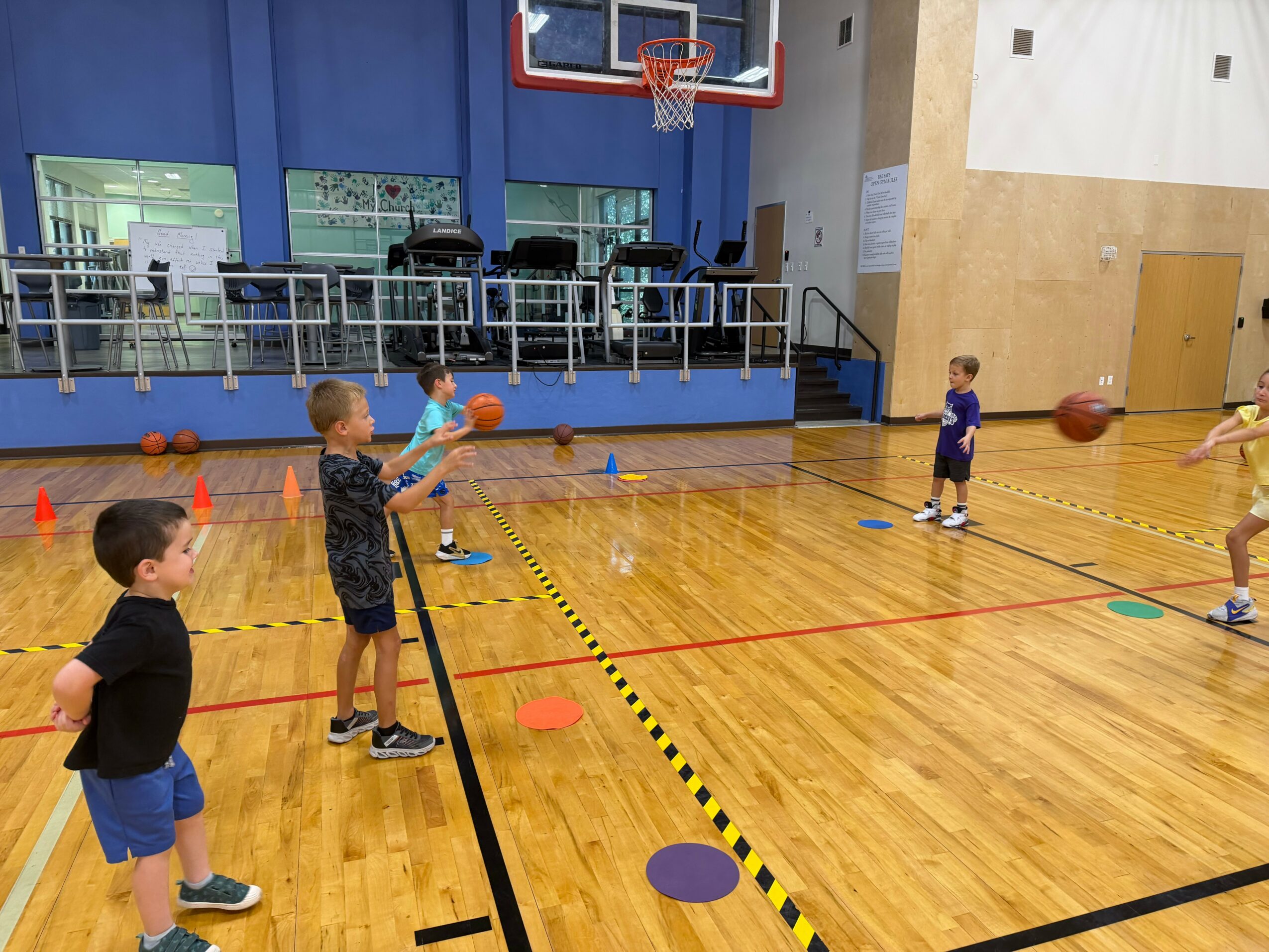 Children playing basketball and practicing dribbling in a gymnasium with wooden floor and blue walls.