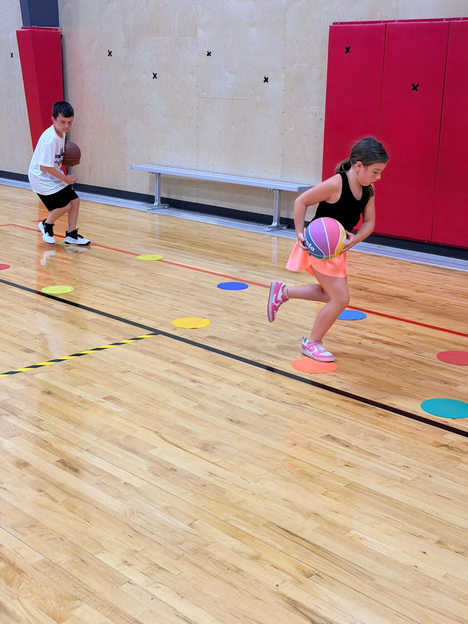 Child in gymnasium holding a ball, standing on colorful markers on wooden floor, with red wall and equipment in background.