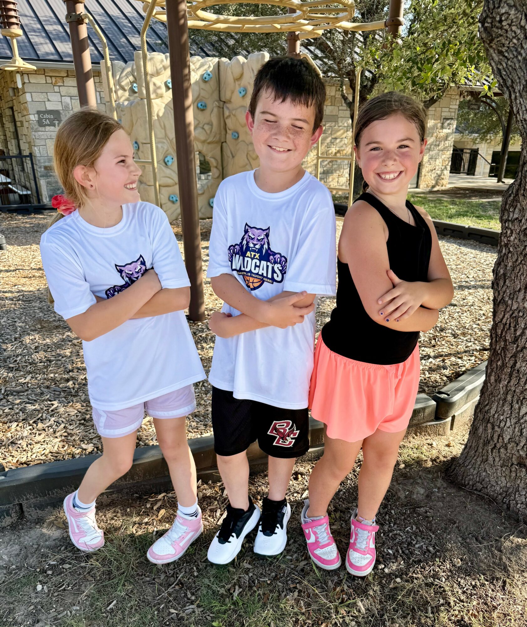 Three children standing outdoors, smiling, with trees and a wooden structure in the background.