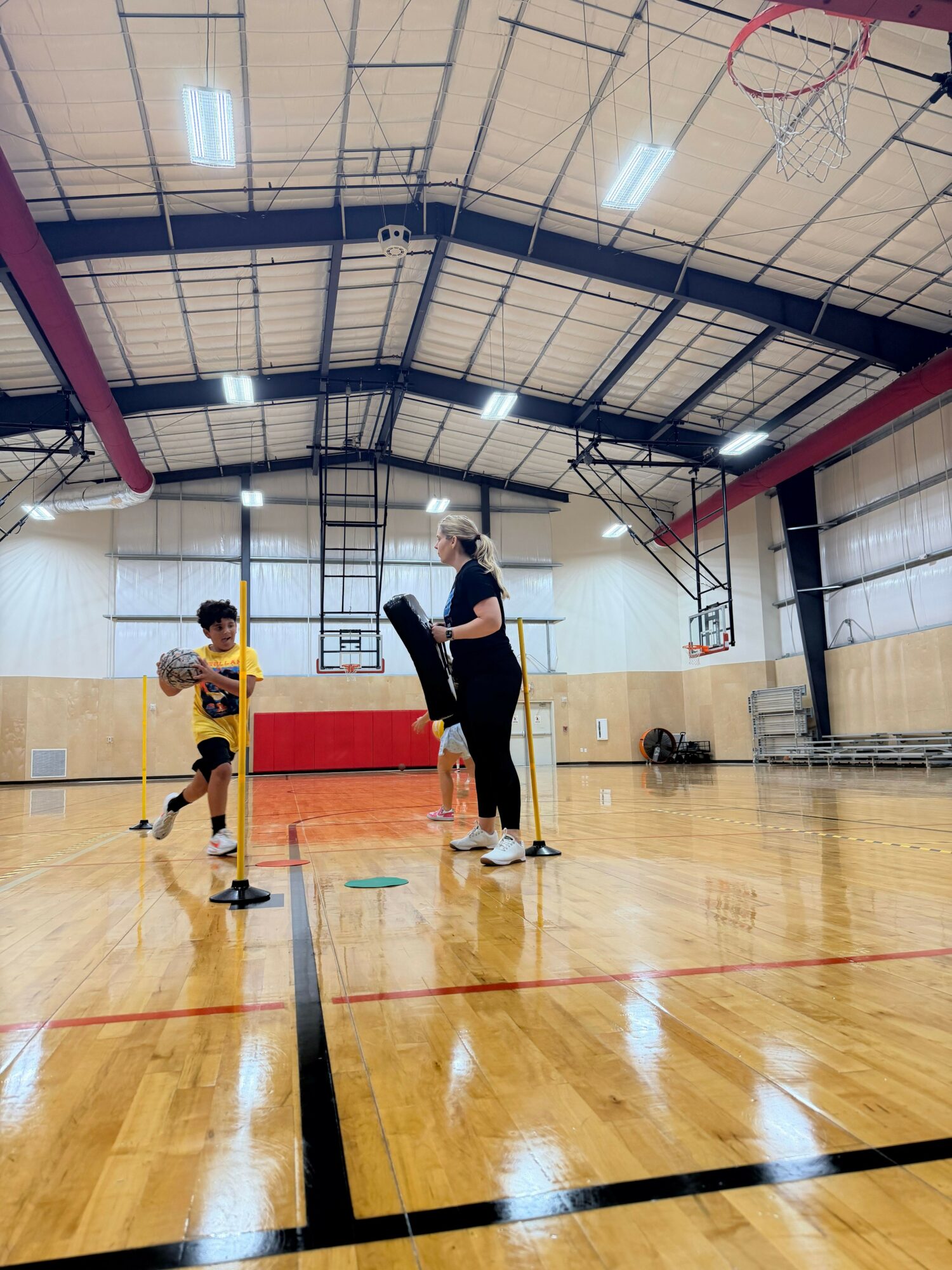 Two children playing basketball in a gymnasium with a basketball hoop and wooden floor.