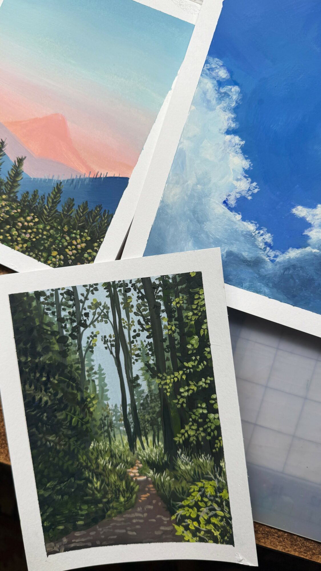 Three landscape photographs of nature scenes, including sky, trees, and a mountain, arranged overlapping on a surface.